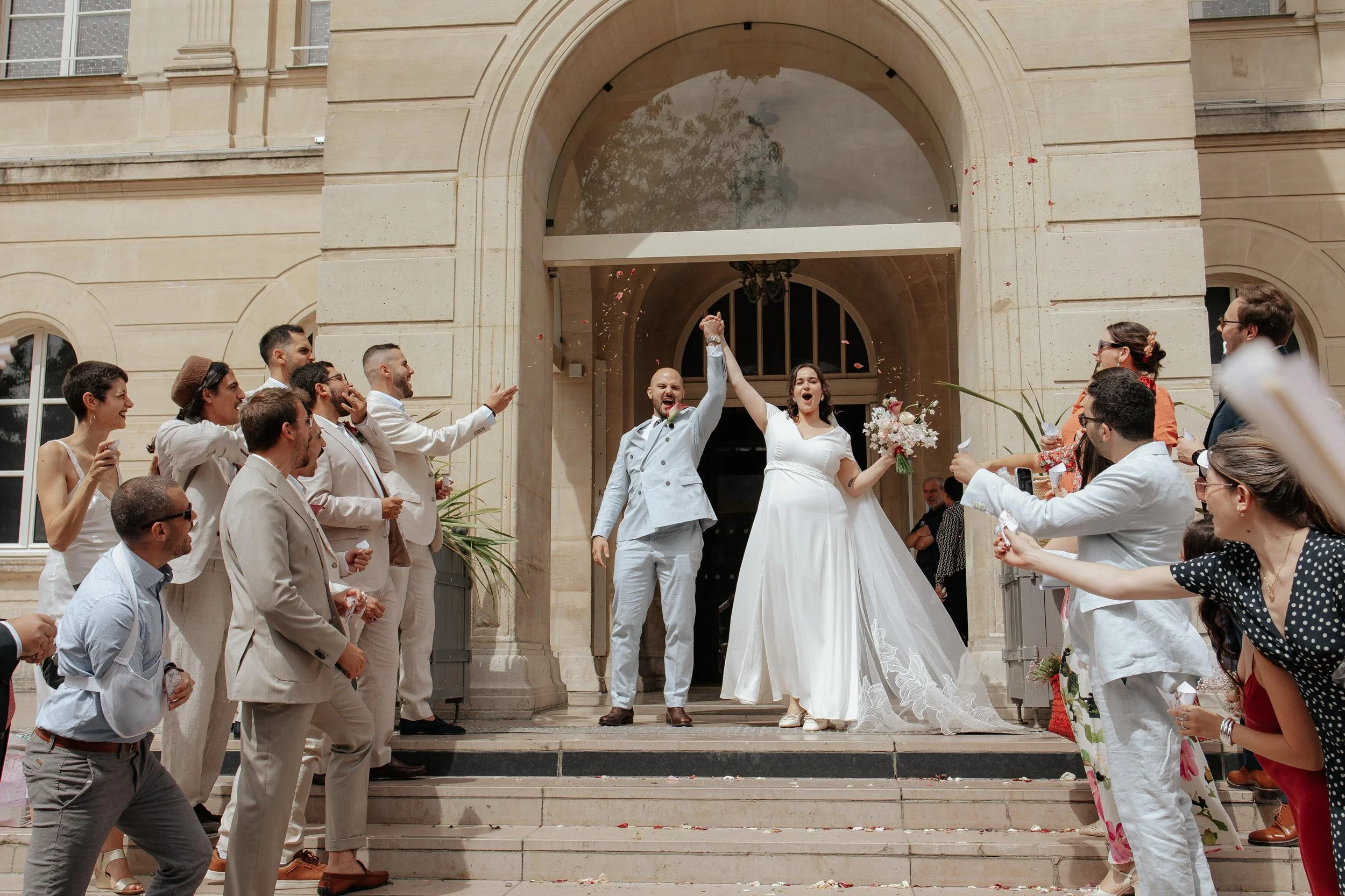 Un couple de mariés joyeux célébrant leur mariage devant un bâtiment avec des invités qui leur lancent des confettis.