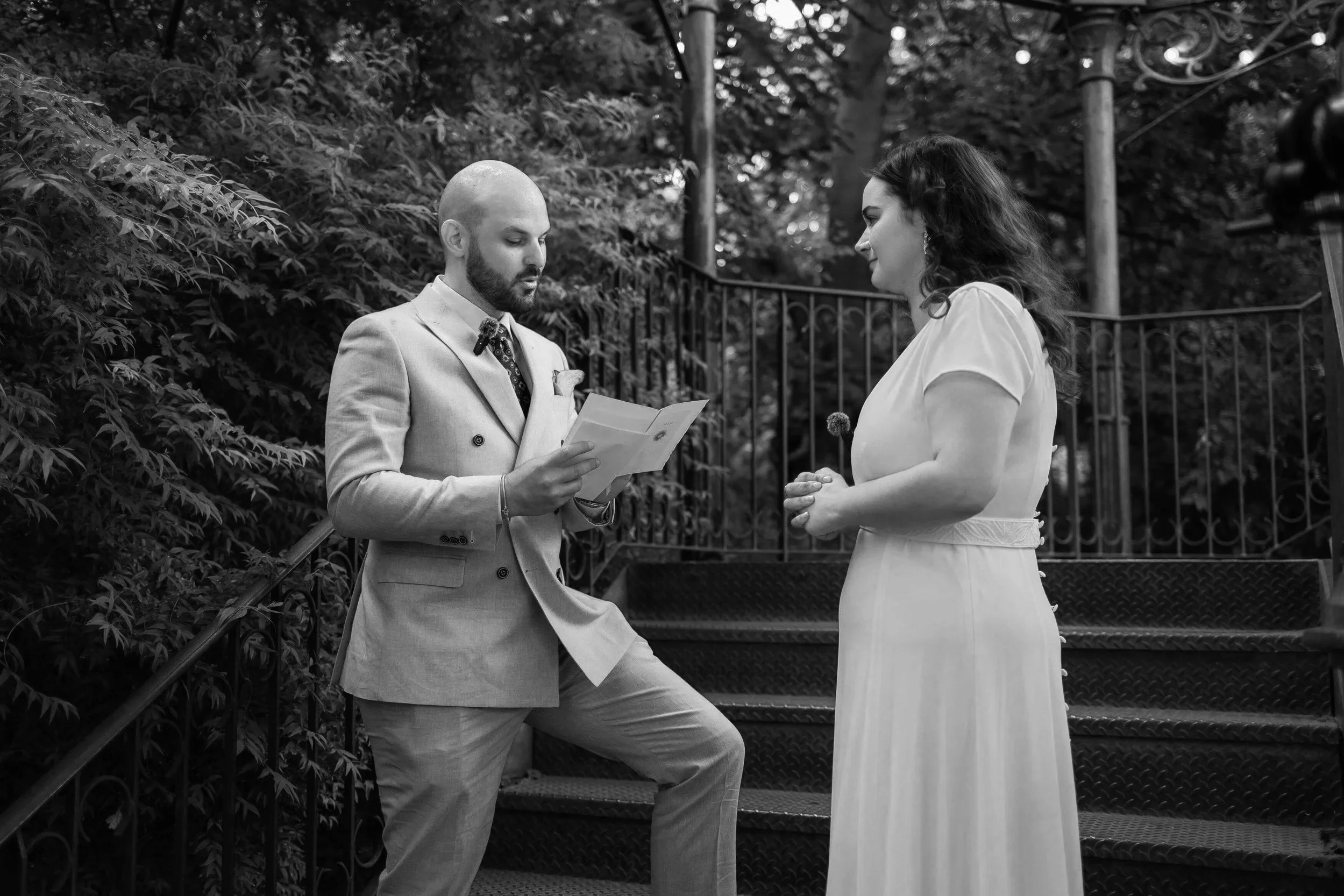 Un homme en costume lit ses vœux à une femme en robe lors d'une cérémonie en extérieur.