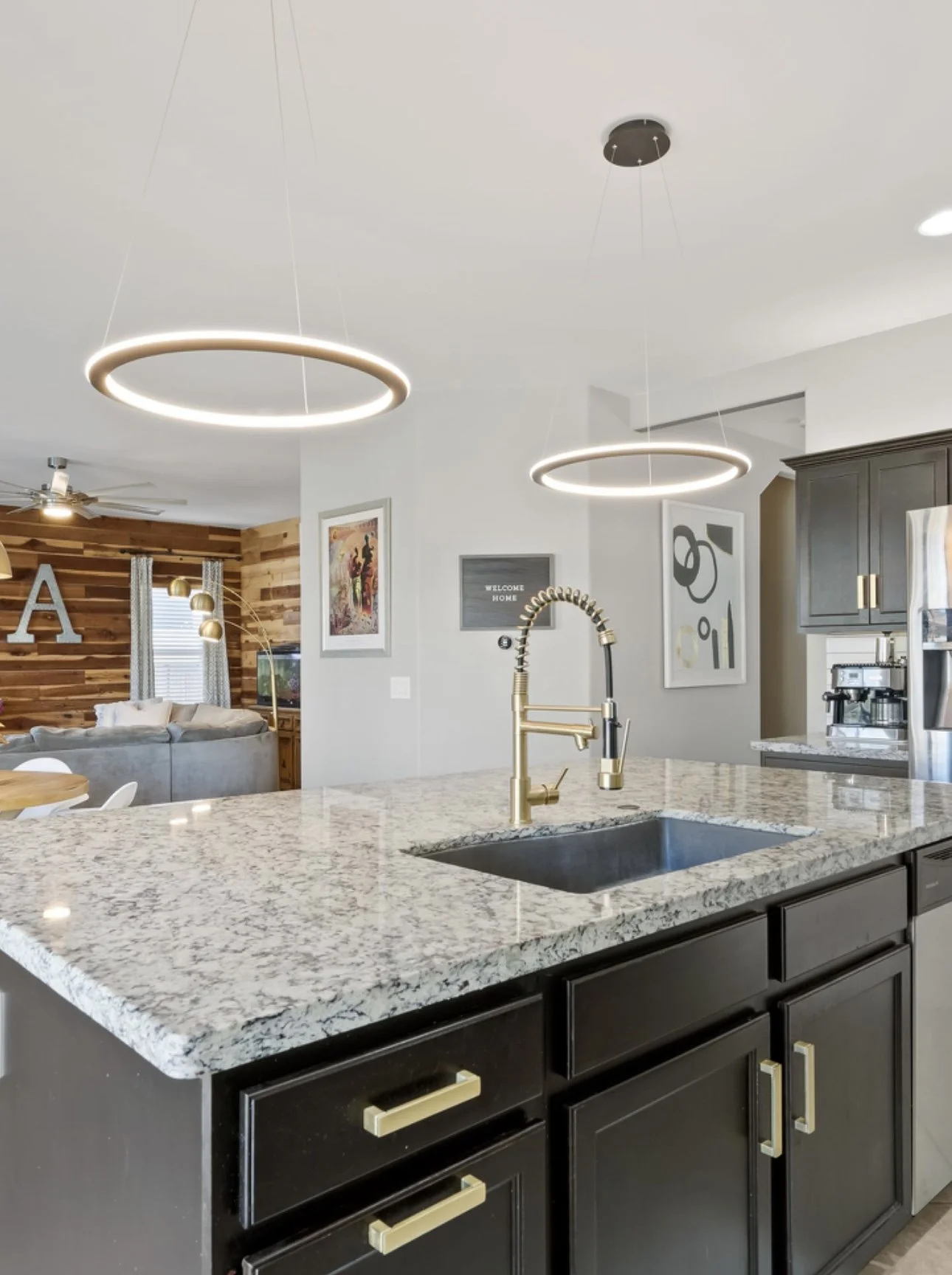 A modern kitchen with a granite island counter, black cabinets with gold handles, a gold faucet, and stylish circular ceiling lights. The background shows a living room with a wooden accent wall and decorative art.