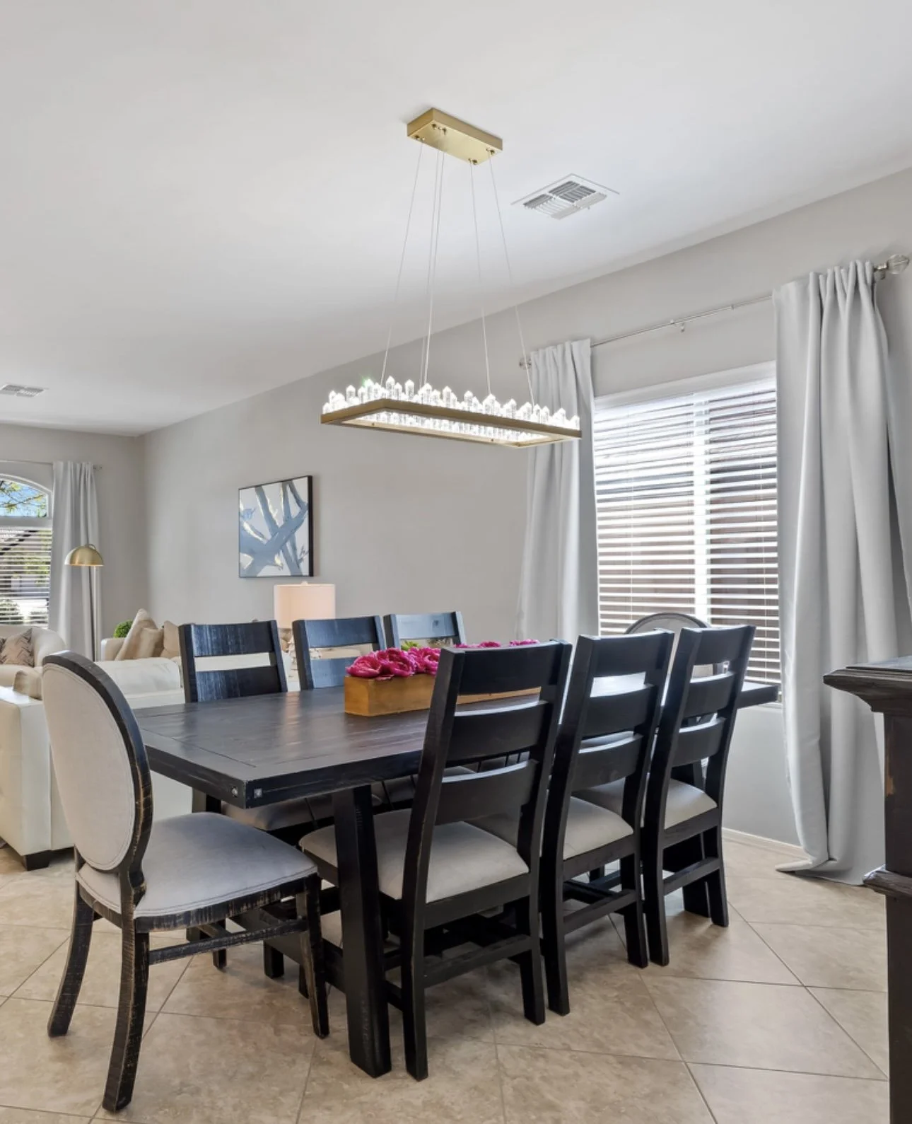 Dining room with black wooden table, seven black chairs, an ornate chandelier, beige tiled floor, and white walls with large windows and white curtains.