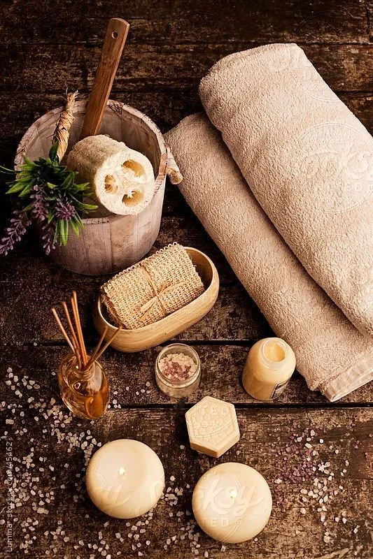 Spa setting with rolled and folded beige towels, candles, bath salts, a loofah, a wooden bucket with spa accessories, a small dish with bath sponge, a glass jar with reed diffuser, and a sprig of lavender with greenery on a dark wooden surface.