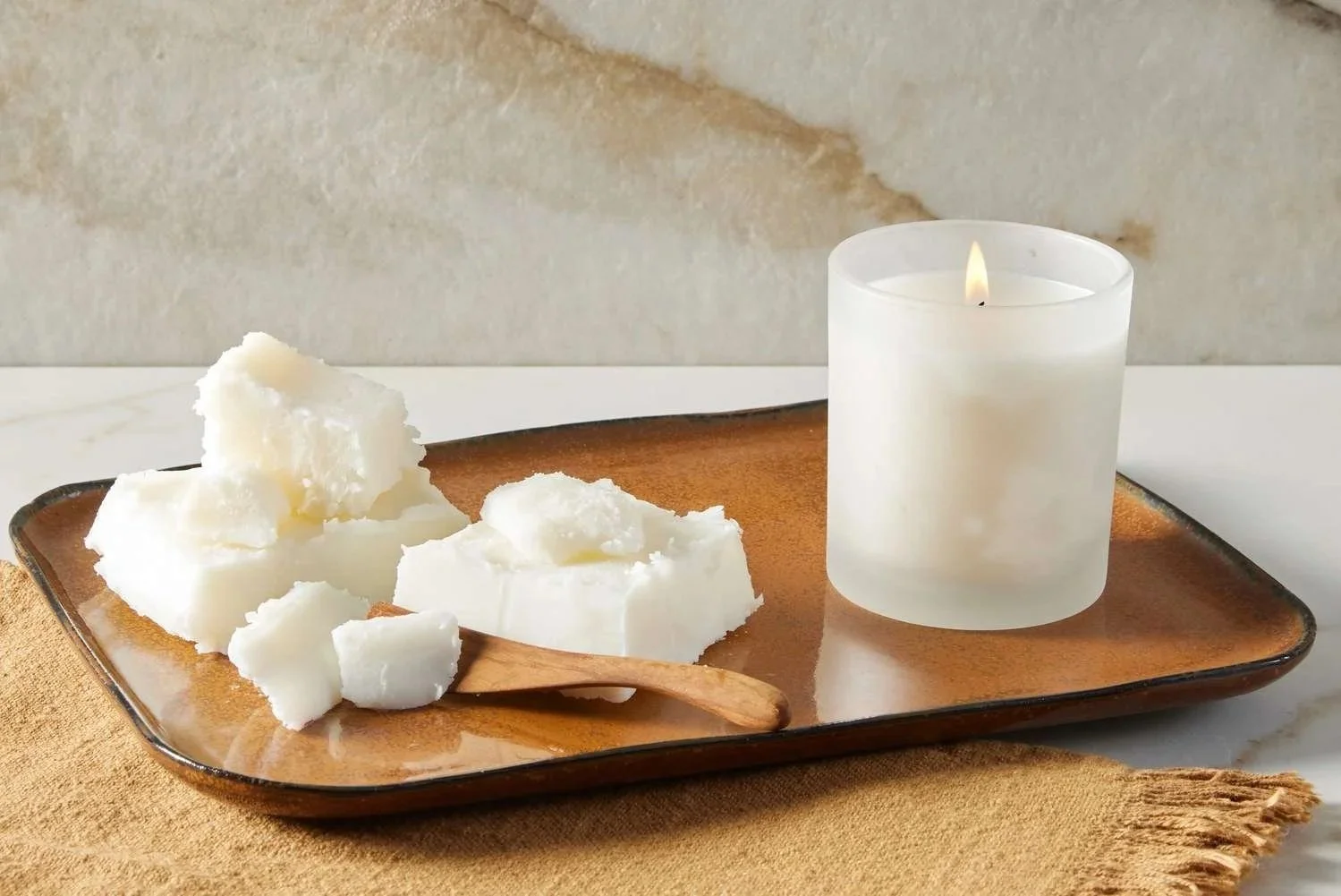 A lit white candle and chunks of shea butter on a brown ceramic tray, placed on a beige cloth.