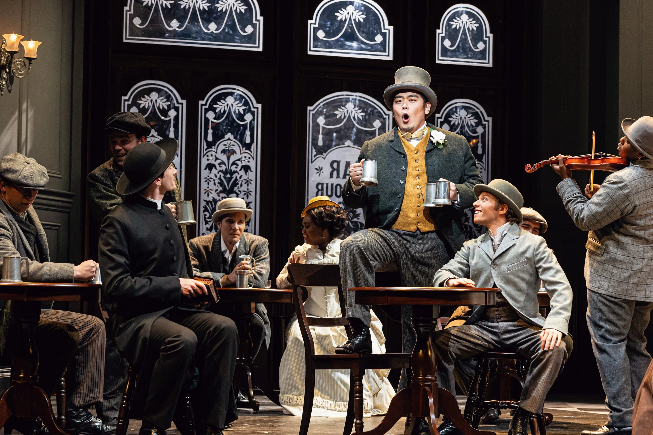 A theatrical performance with actors in vintage attire, some seated at tables and one standing on a chair holding mugs, others playing music and engaging in lively conversation.