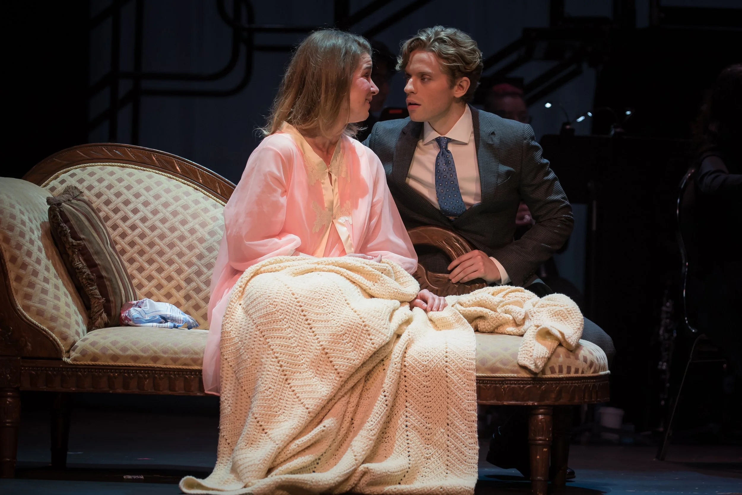 A scene from a theatrical play showing a woman in pink pajamas and a man in a suit sitting closely on a vintage sofa, engaged in an intimate conversation.