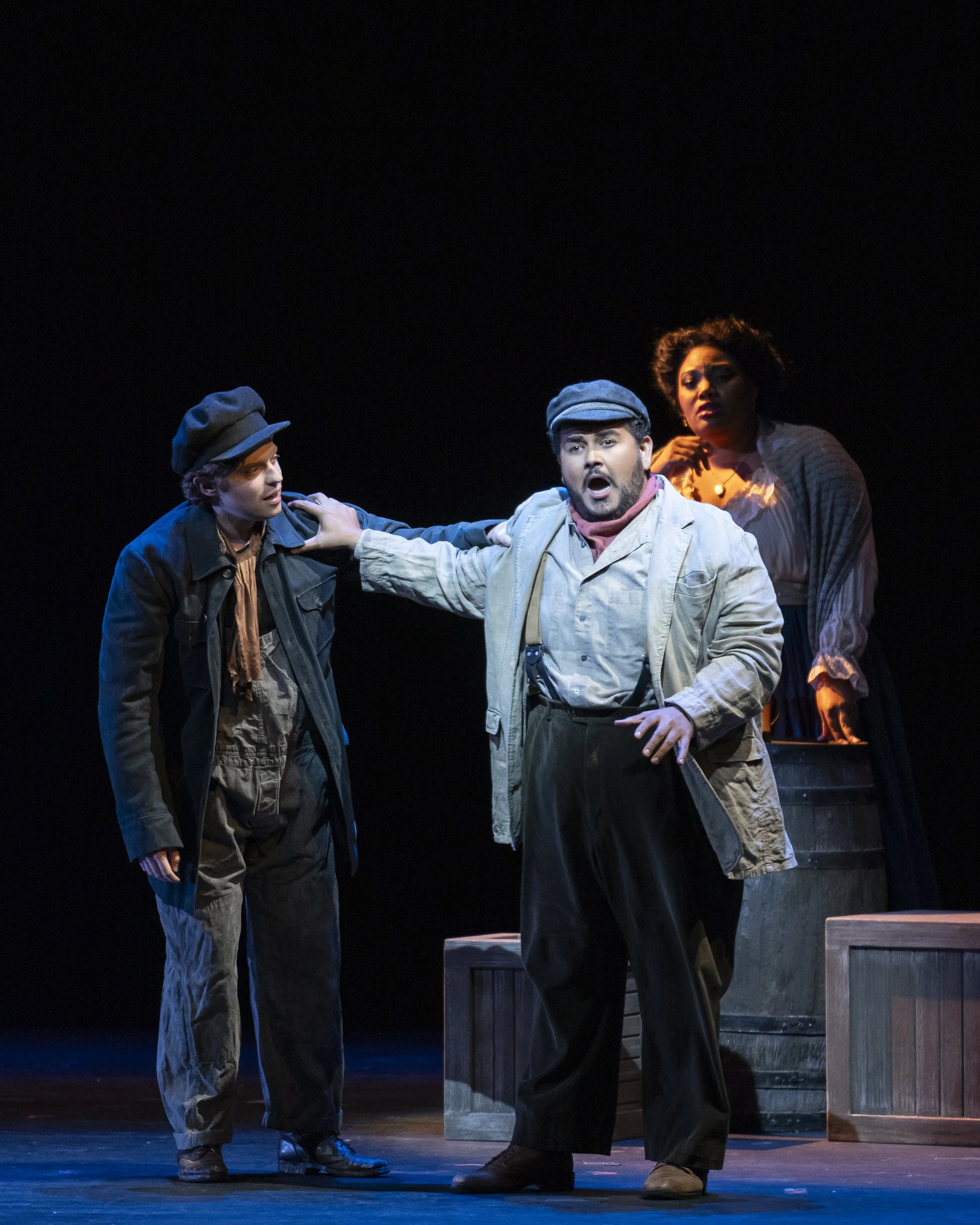 Three actors perform on stage with a dark background; one man is in the center, gesturing with an expressive facial expression, while a woman stands behind him, and another young man is to his left, listening.