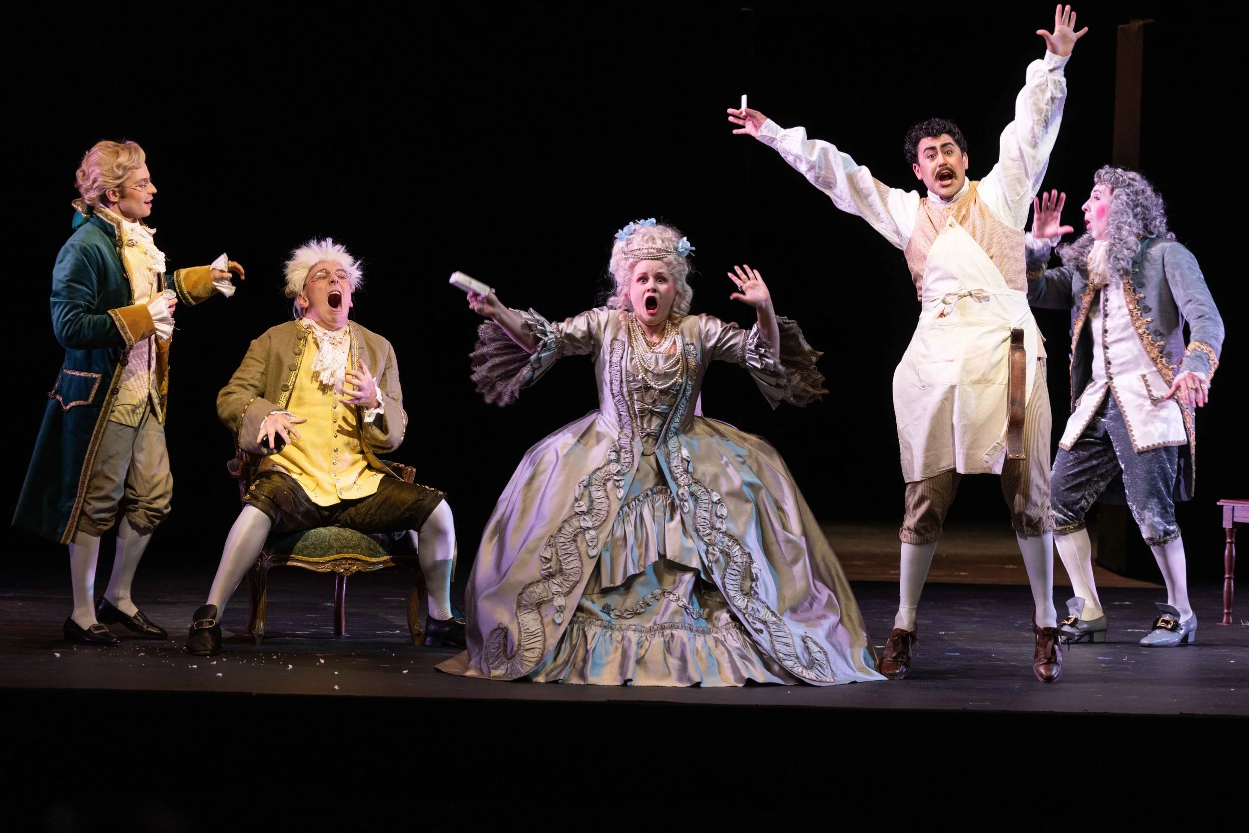 Actors performing on stage in period costume, with one woman in a large, elaborate gown, appearing distressed. Four men and women around her, all in historic attire, showing surprise or concern.