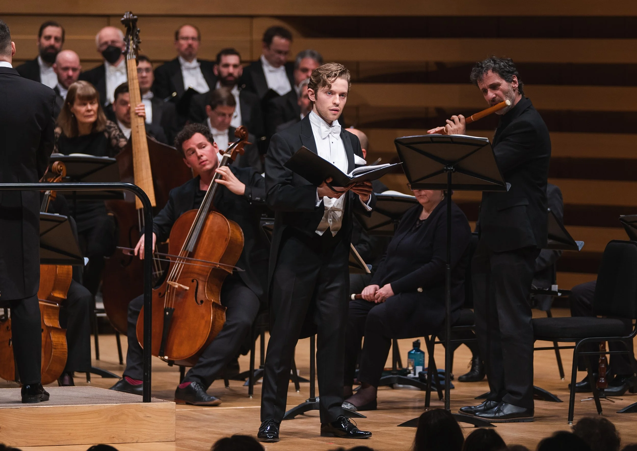 Orchestra performing on stage with musicians in formal attire, using music stands, and a wooden backdrop.
