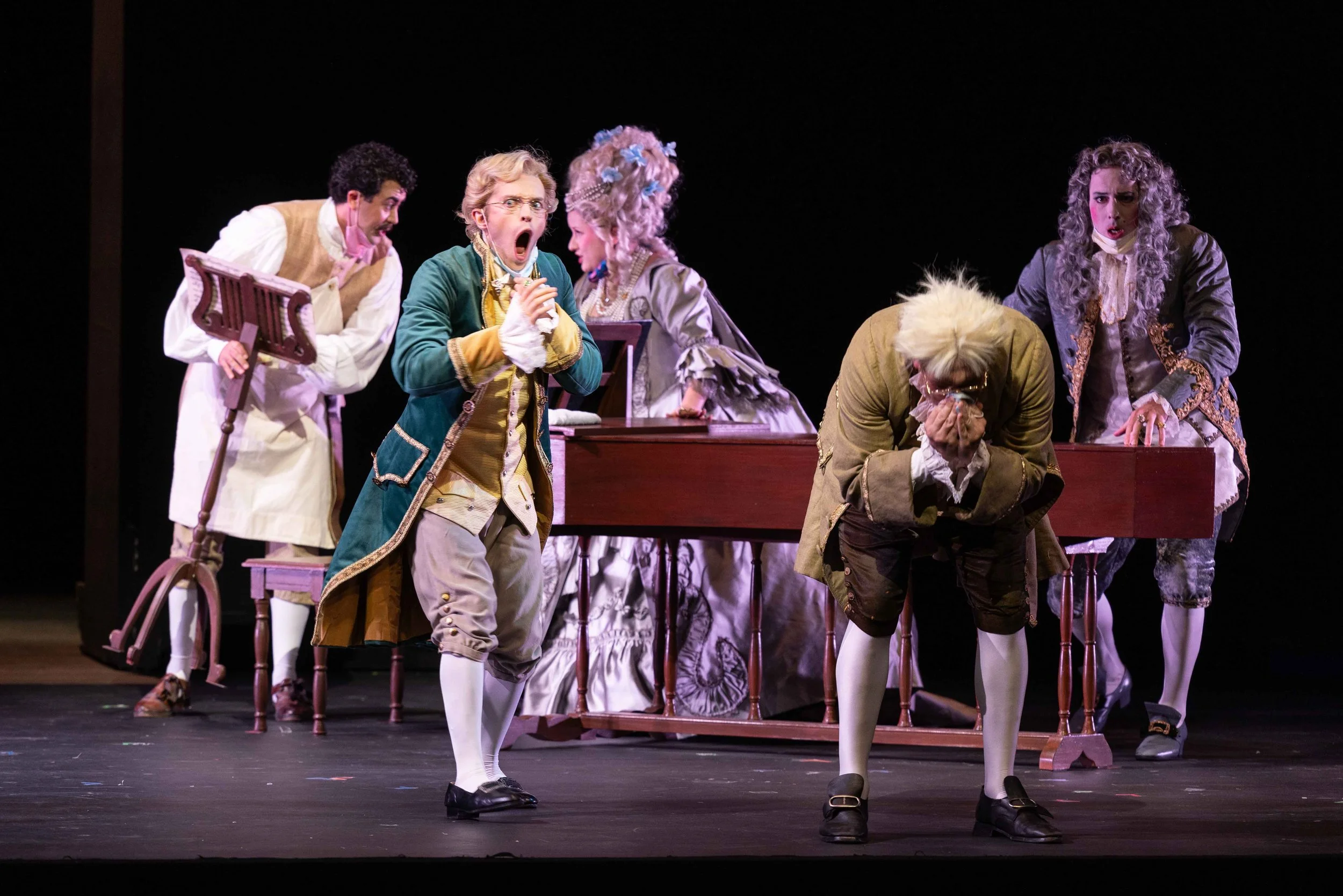 Actors in 18th-century costumes perform a theatrical scene on stage, with expressions of shock and surprise.