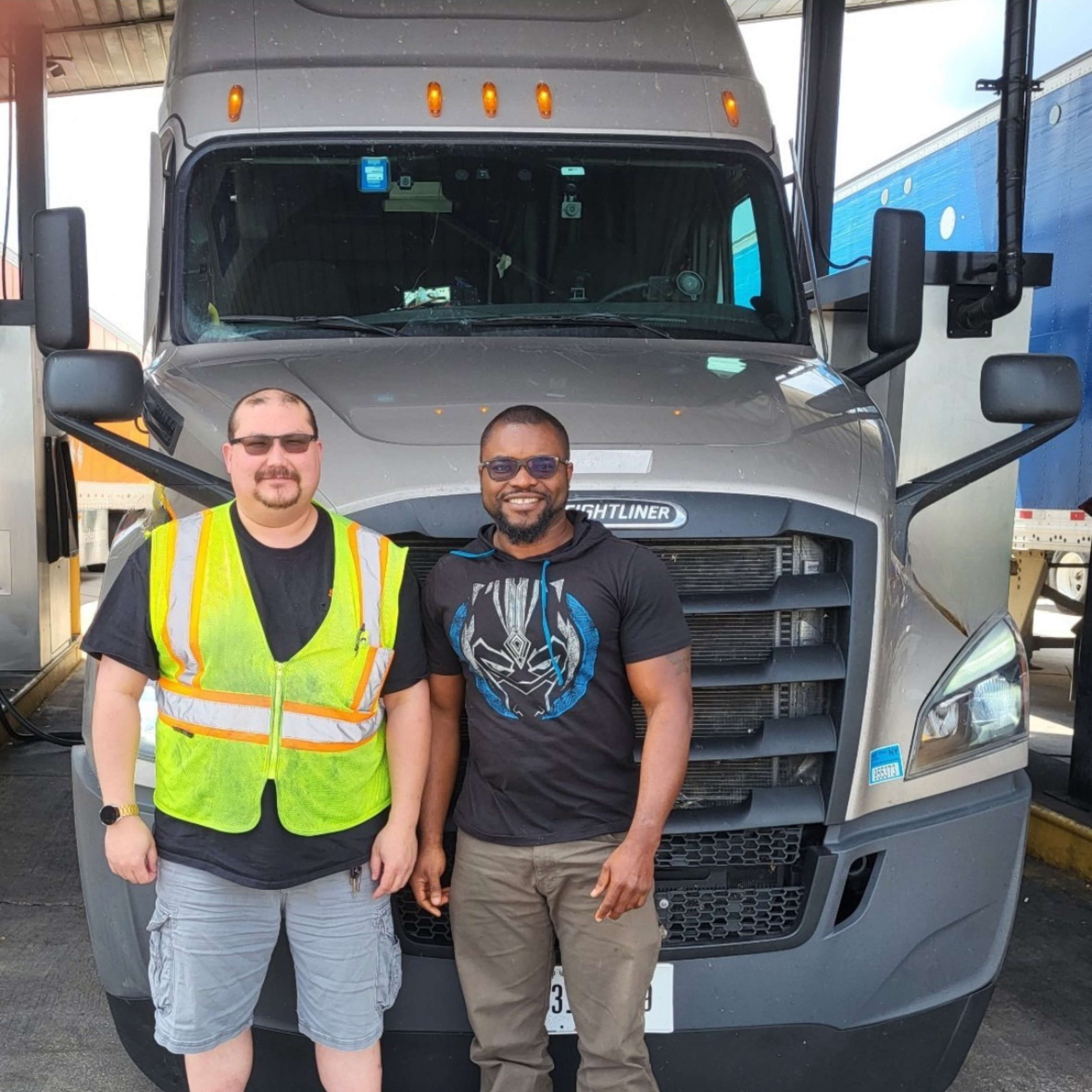 Two men standing in front of a large semi-truck, smiling at the camera. One wearing a black t-shirt with a graphic and a black hoodie, the other wearing a black shirt and a yellow safety vest.