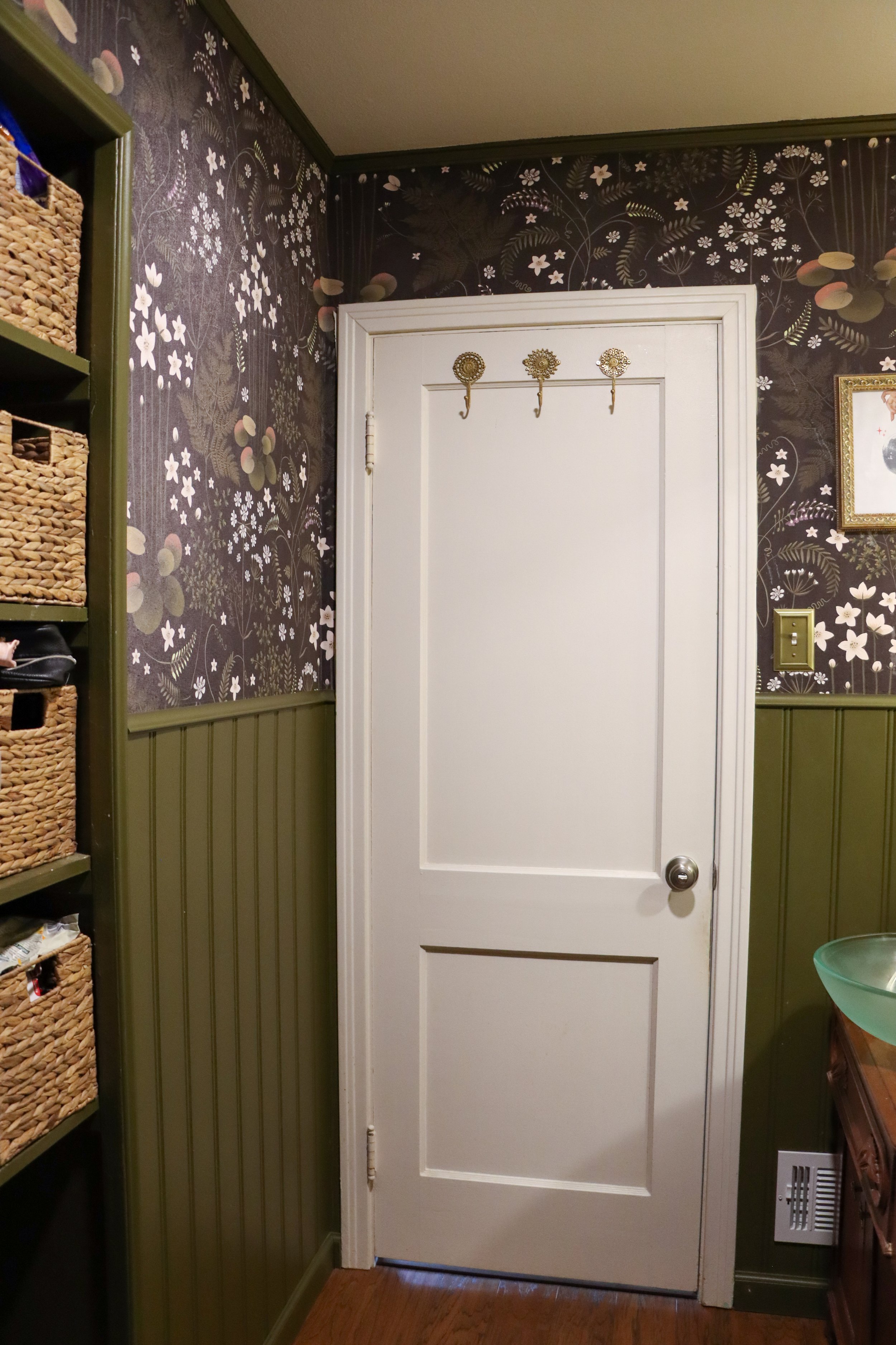 A white interior door with three decorative hooks at the top, set in a room with dark floral wallpaper and green wainscoting, next to a set of woven baskets on a green shelf, a framed picture on the wall, and a glass bowl on a wooden surface.