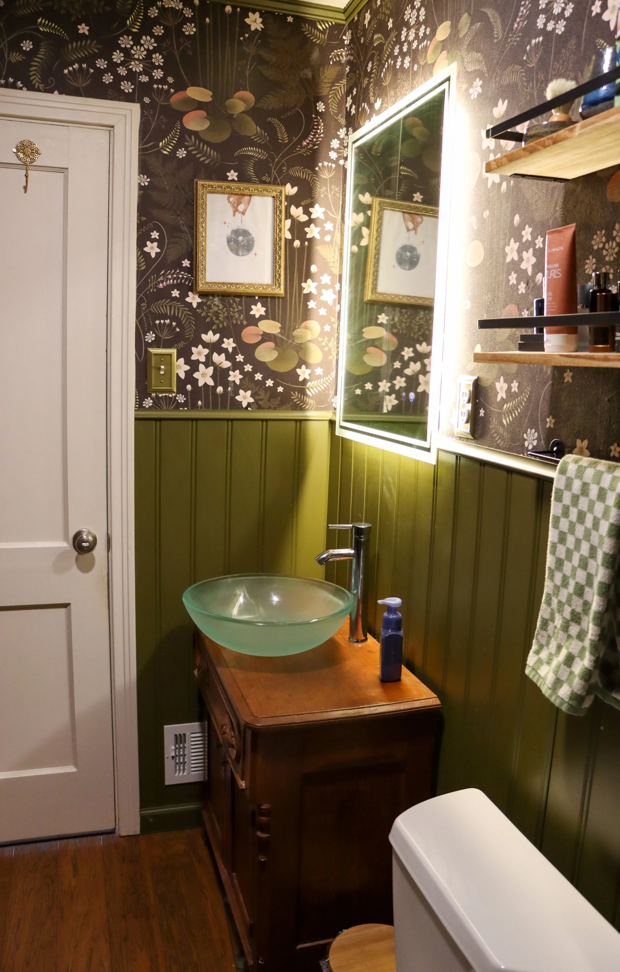 A small bathroom with a floral wallpapered wall, green wooden paneling, a mirror with LED lighting, and a vessel sink on a vintage wooden cabinet. There are shelves with toiletries and a towel hanging, with a toilet partially visible in the foreground.