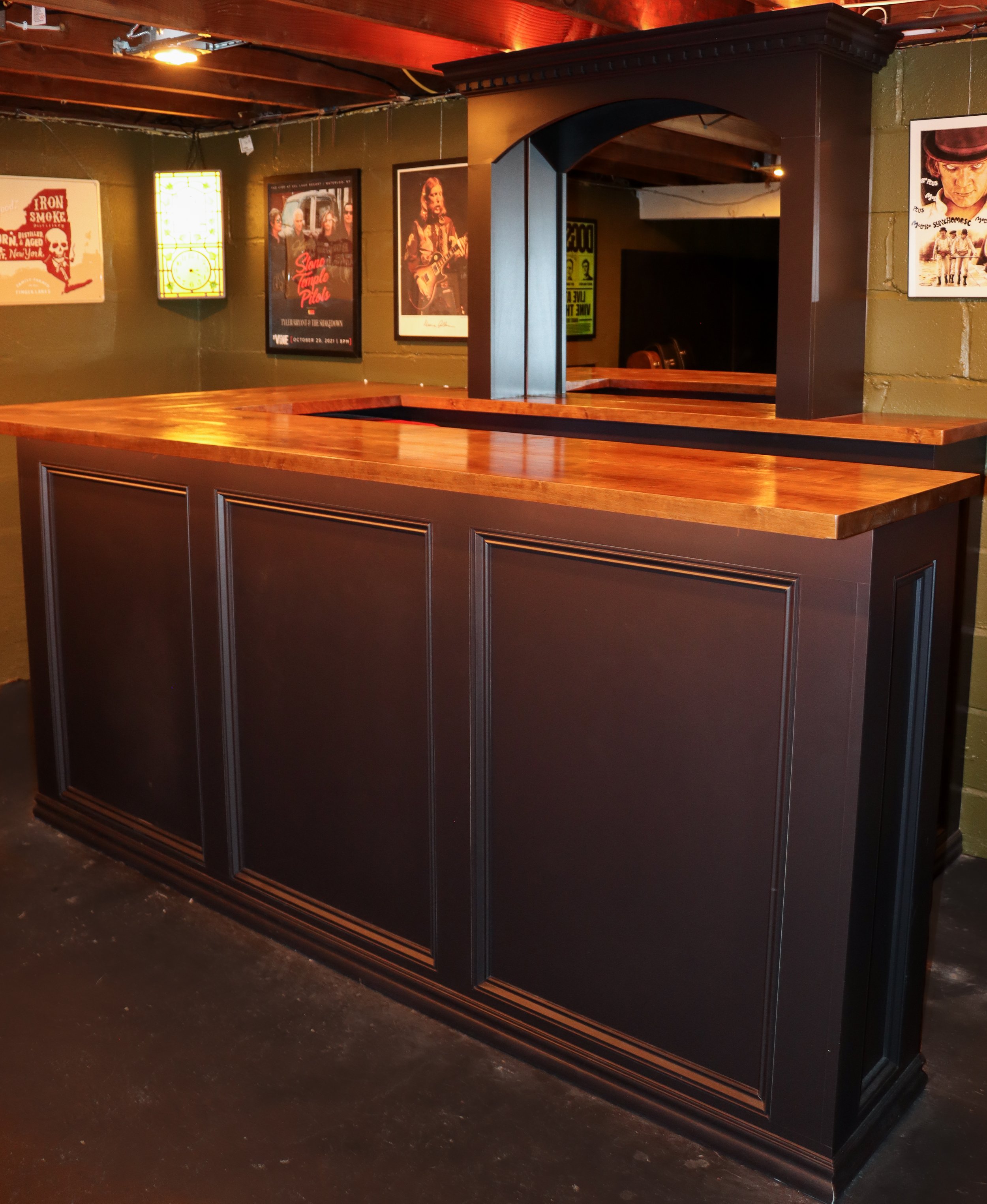 View of a dark wooden bar counter with a polished wood top and decorative framing, set in a room with vintage movie posters and wall art.