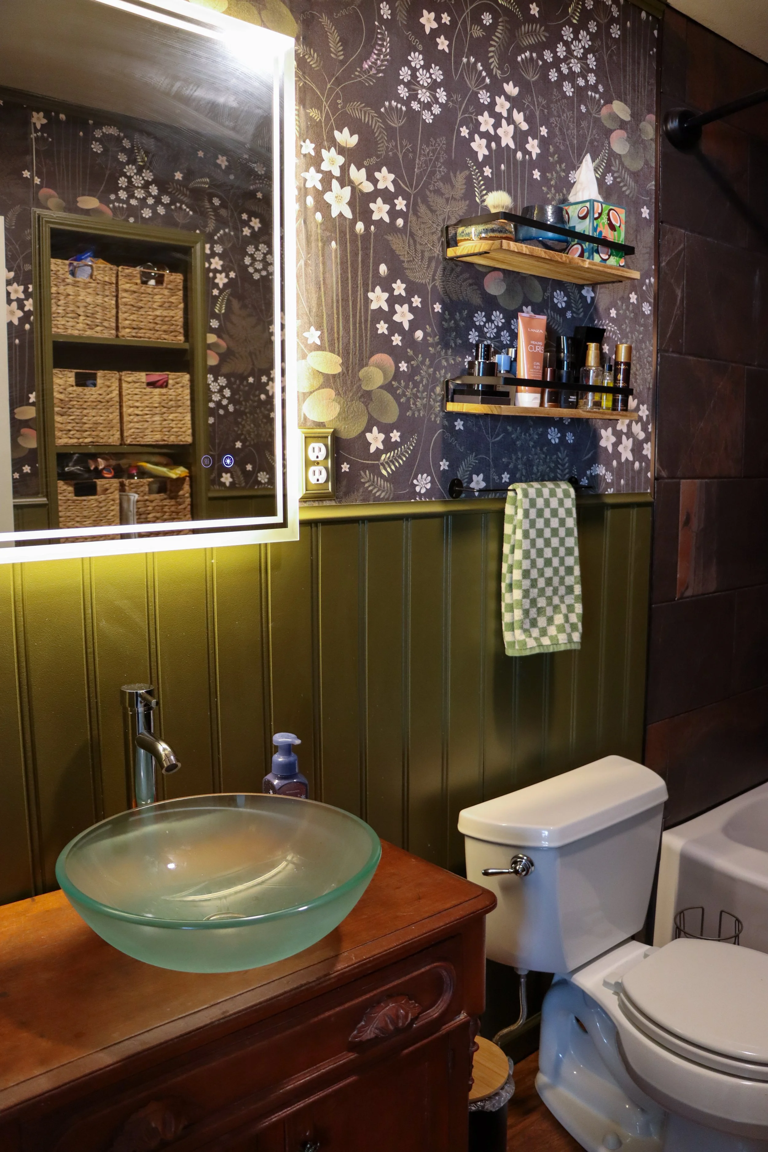 A bathroom with a decorative mirror, floating shelves with toiletries, a glass vessel sink on a wooden vanity, a toilet, and a wall featuring floral wallpaper and green wainscoting.
