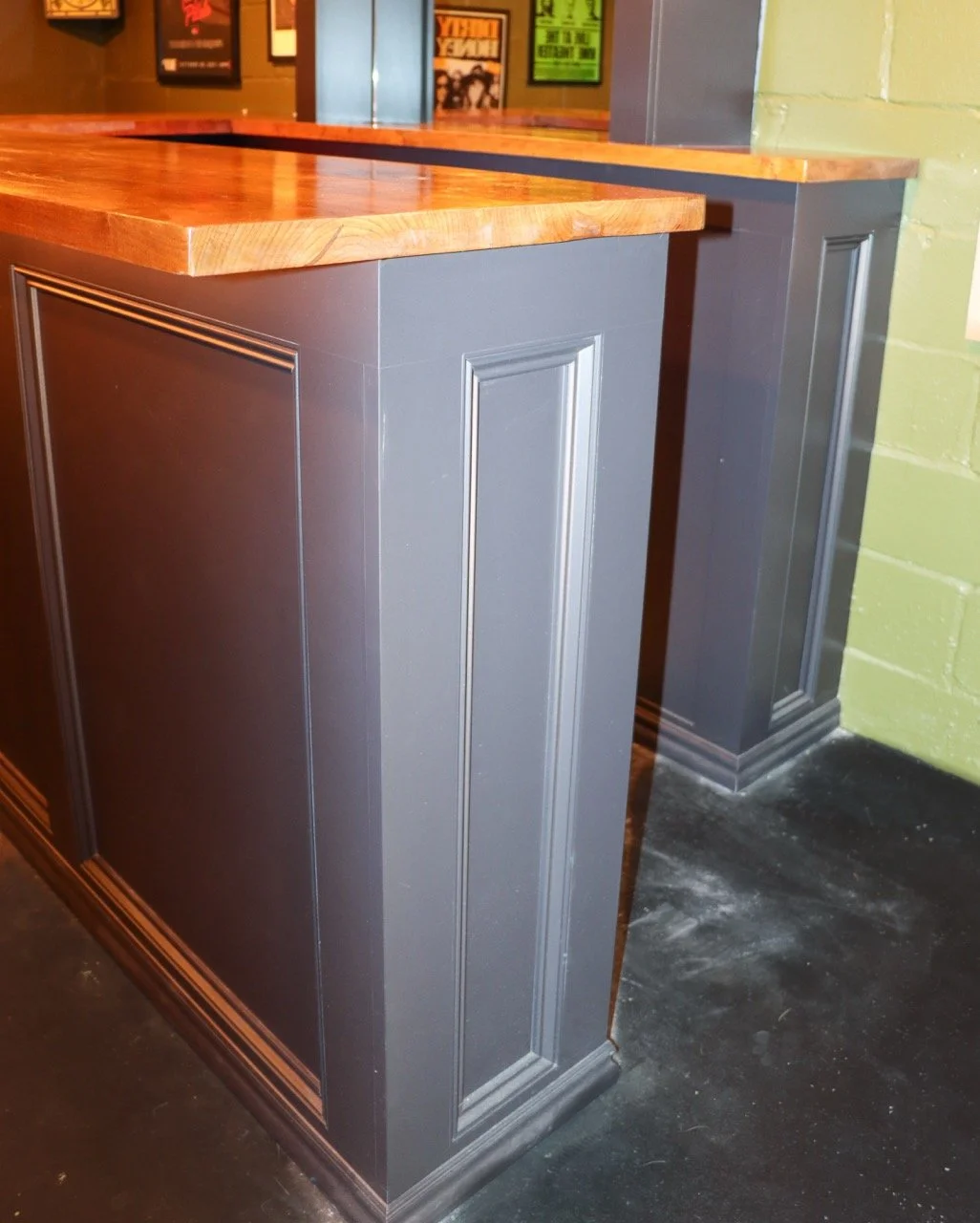 Close-up of a bar counter with a dark gray paneled base and a polished wooden countertop, in a dimly lit venue decorated with colorful framed posters on the wall.