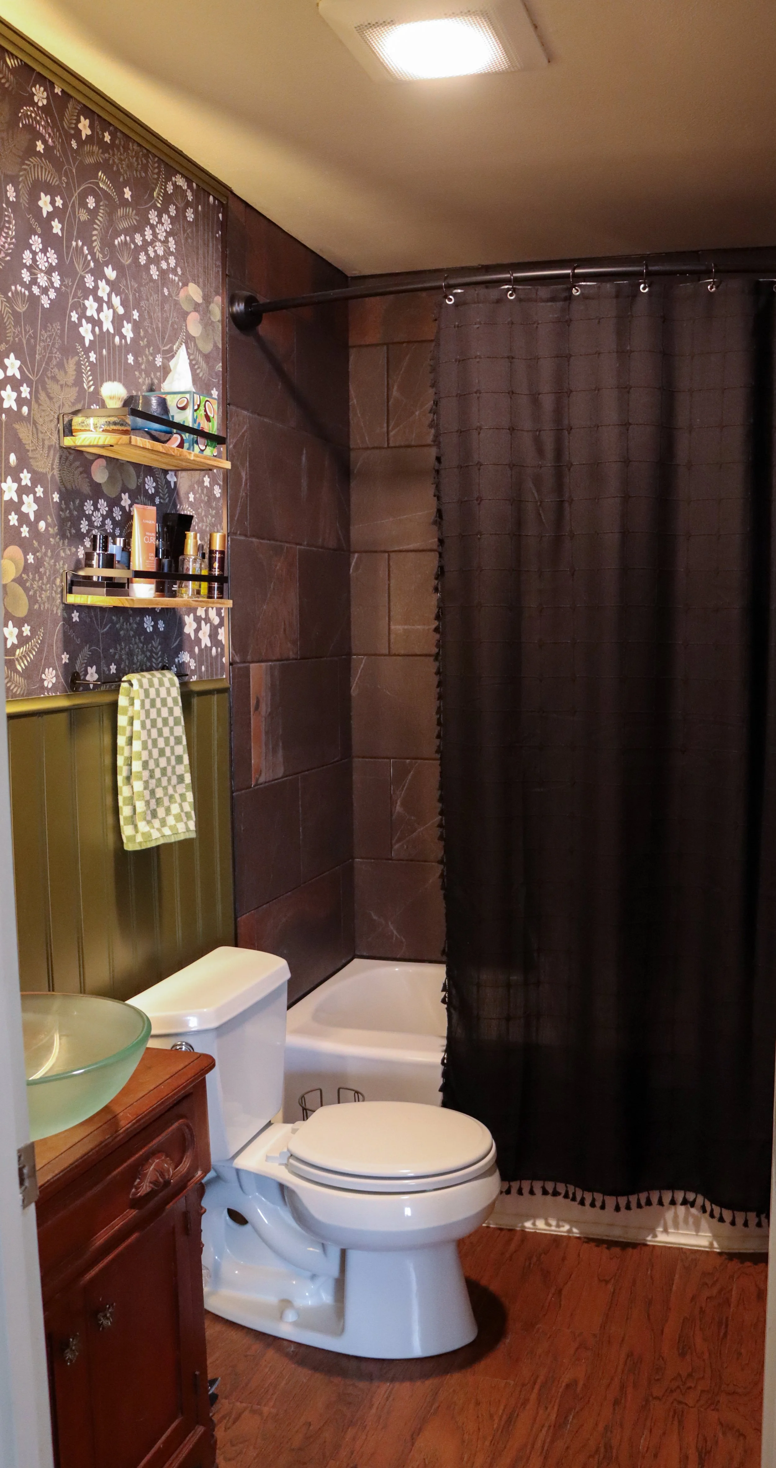 A small bathroom with a toilet, a wooden vanity with a glass vessel sink, a patterned wallpaper, dark brown tiled walls, a black shower curtain, and a ceiling light.