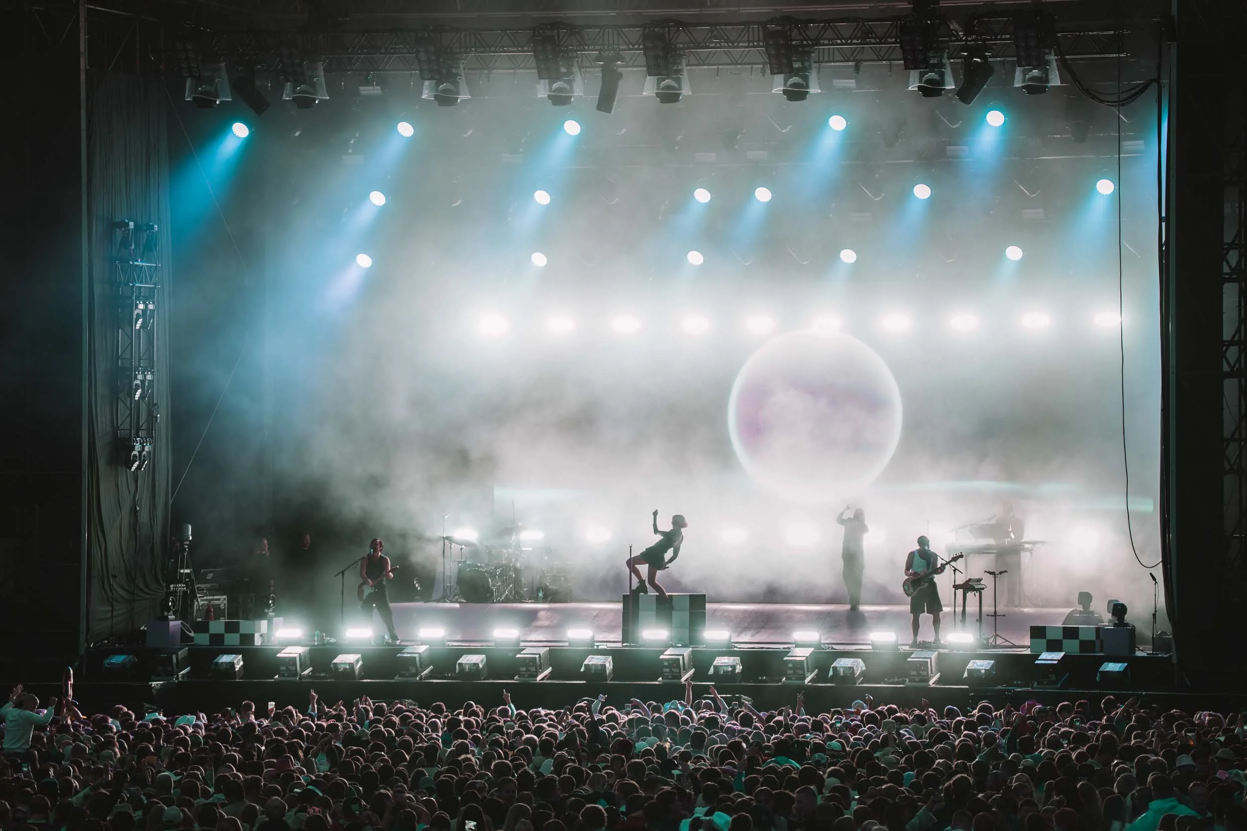 En scene med et band som spiller under en konsert, med lys og tåke, og en stor folkefest foran scenen.