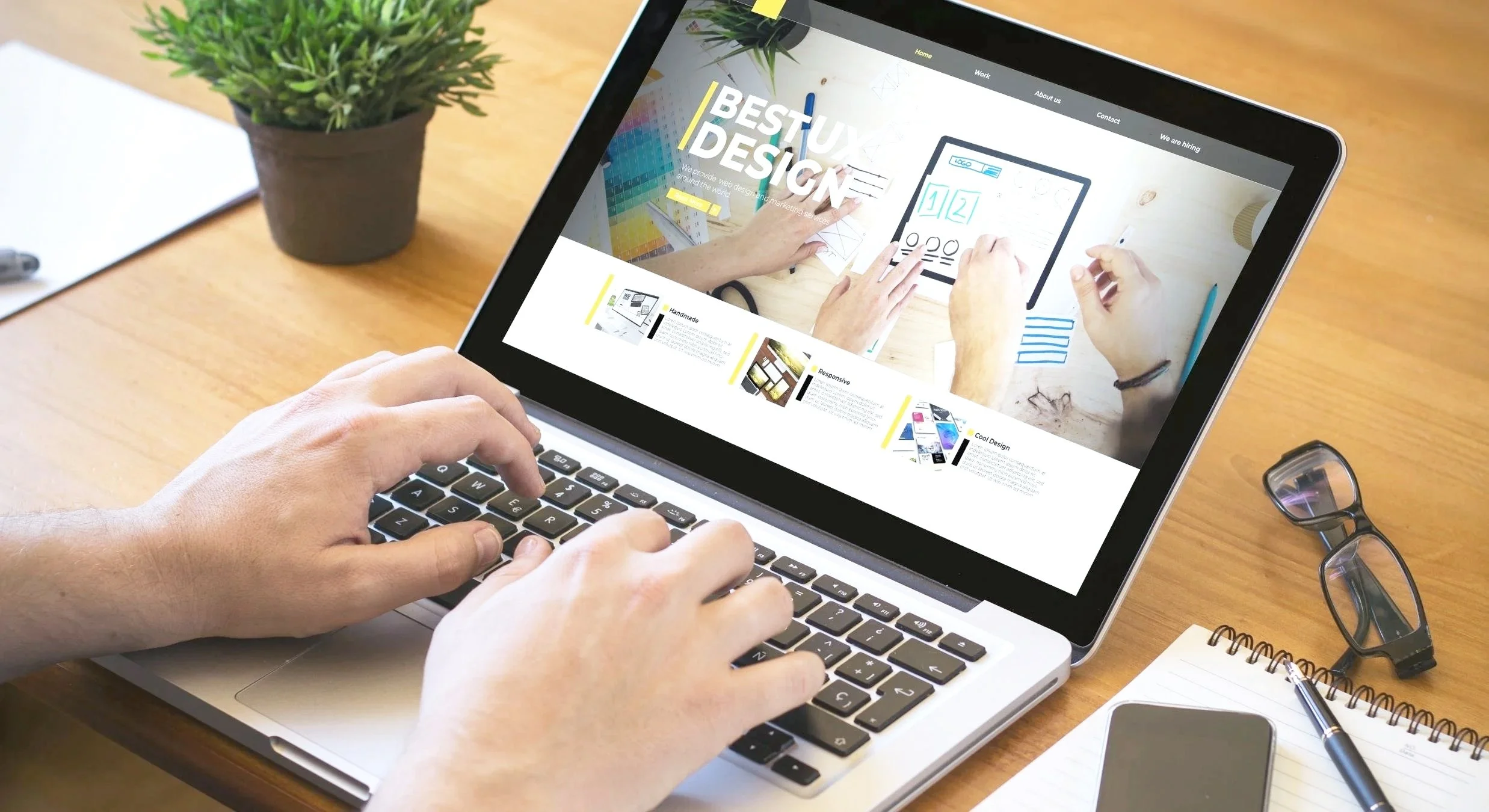 En person bruker en bærbar datamaskin på et trebord, med en dørklokke, notatblokk, penn, smarttelefon, lesebriller og en pottesplante rundt.