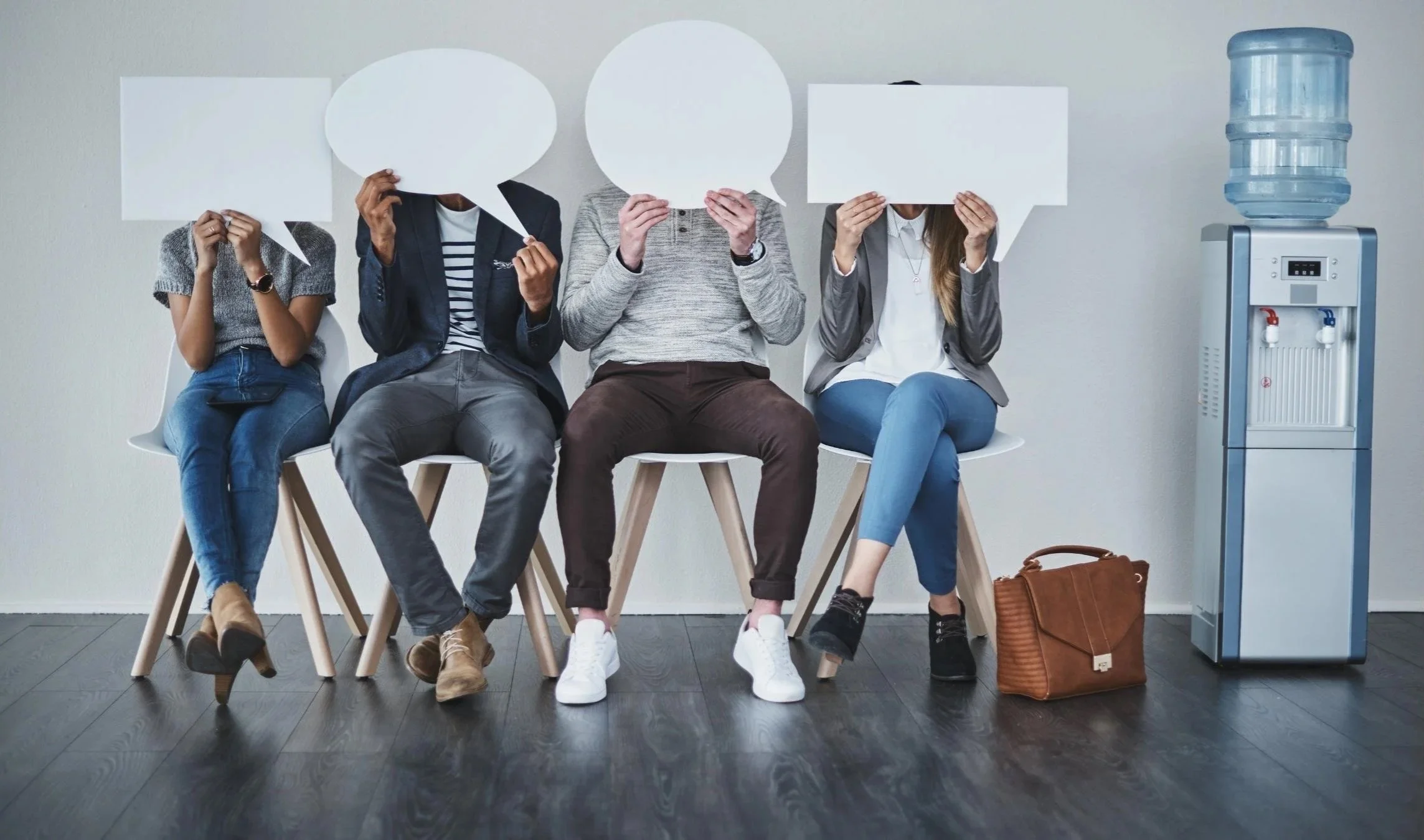 Fire personer sitter på stoler mot en hvit vegg, holder opp snakkebobler som dekker ansiktene sine, og det står en vannkjøler til høyre.