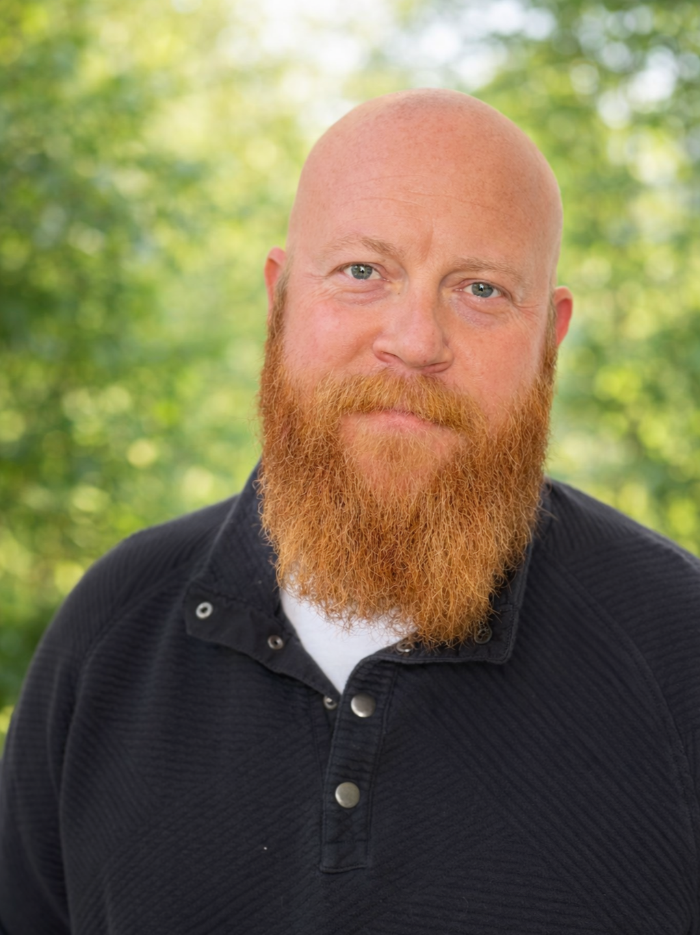A man with a bald head, blue eyes, and a full red beard looking at the camera, outdoors with a green blurred background.