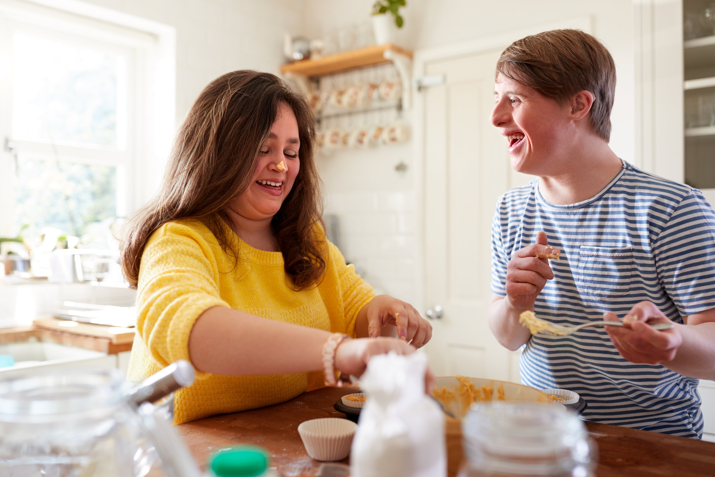Two people, a woman in a yellow sweater and a young man in a striped shirt, are laughing and baking together in a bright kitchen. The woman is smiling and has a piece of dough on her nose, while the man holds a fork with dough on it, both showing joy.