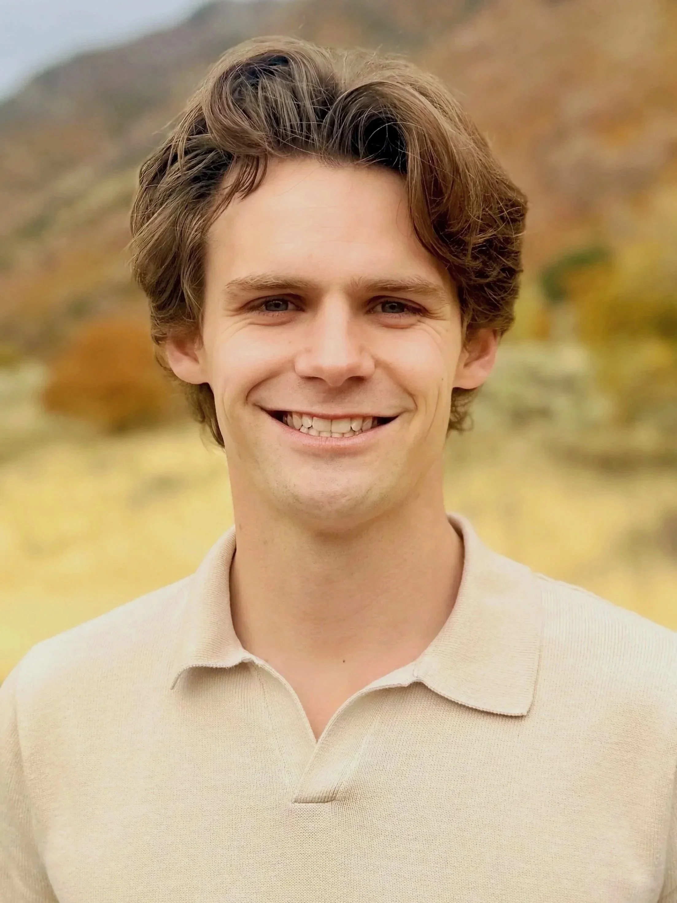 A young man with brown wavy hair smiling outdoors during autumn, wearing a beige polo shirt, with trees and mountains in the background.
