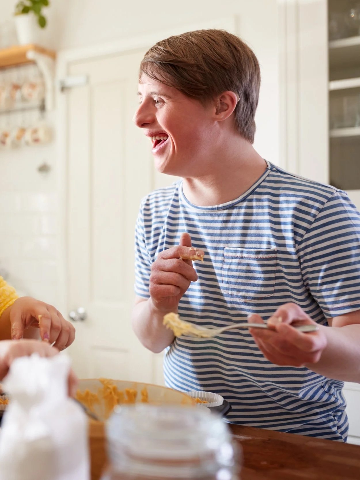 A young man with light skin and brown hair, wearing a blue and white striped shirt, smiling while holding a fork with food, in a kitchen setting.