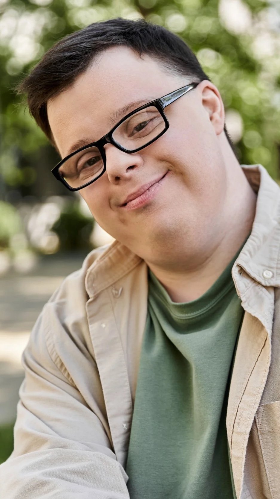 A young man with short dark hair, glasses, and light skin, smiling outdoors in a park-like setting, wearing a beige jacket over a green shirt.