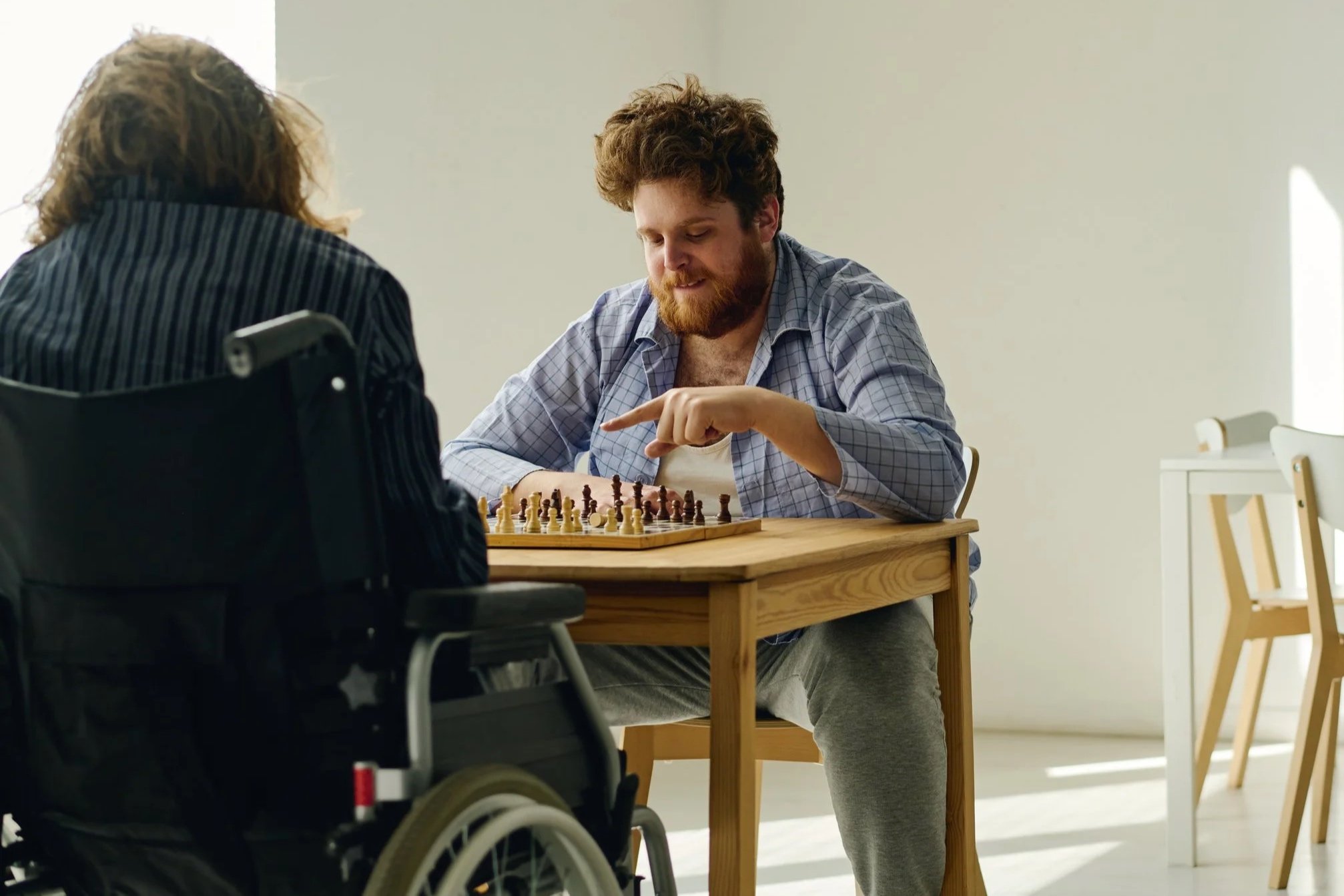 Two men playing chess indoors, one in a wheelchair, the other sitting across the table, with bright natural light coming through a window.