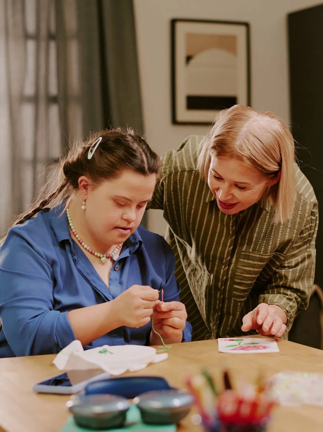 A girl with a colorful beaded necklace working on a craft project at a table, being guided by a woman with blonde hair leaning over her.