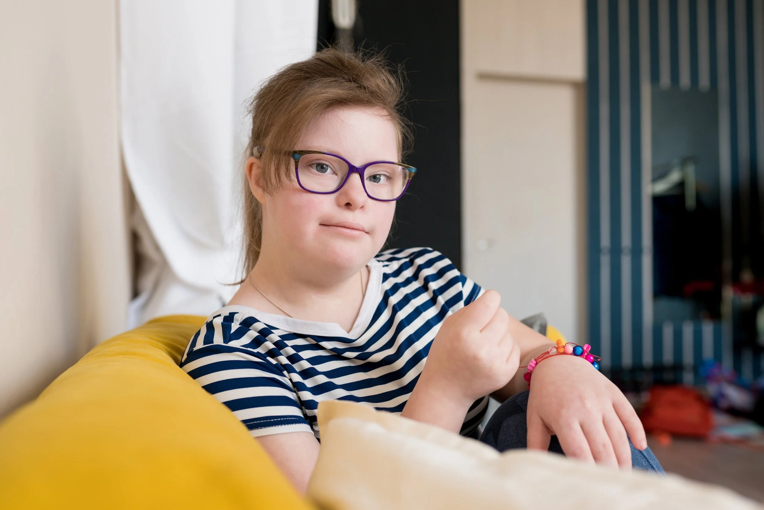 Young girl with glasses in a striped shirt sitting on a yellow couch, looking at the camera, showing a wrist with colorful beads, in a cozy room.