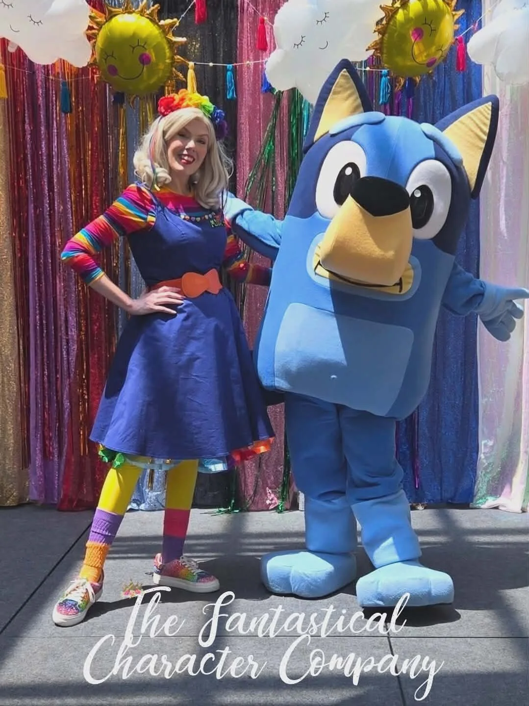 A woman dressed in a colorful rainbow-themed outfit poses next to a person in a blue cartoon character costume, both standing in front of a vibrant, rainbow-colored backdrop with balloons and clouds. The image includes the text "The Fantastical Character Company."