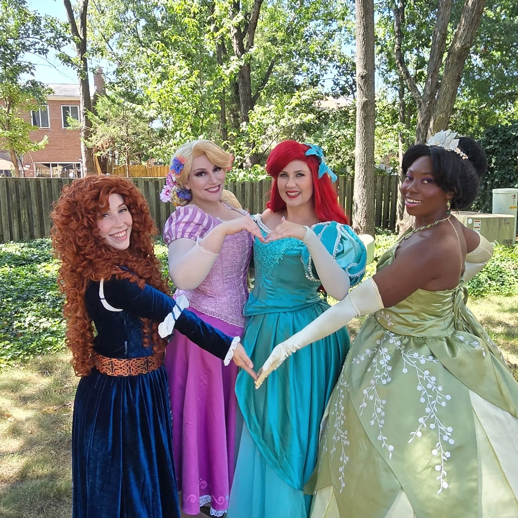 Four women dressed as Disney princesses posing together outdoors under trees.