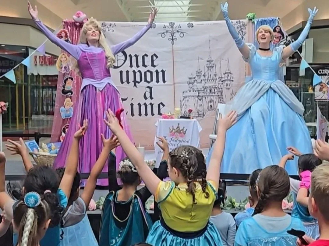Children watching a stage performance with two women dressed as Disney princesses, Rapunzel in purple and Cinderella in blue, in a shopping mall.