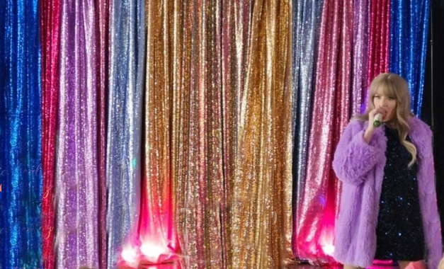 A woman singing on stage in front of a colorful, sparkly curtain backdrop.