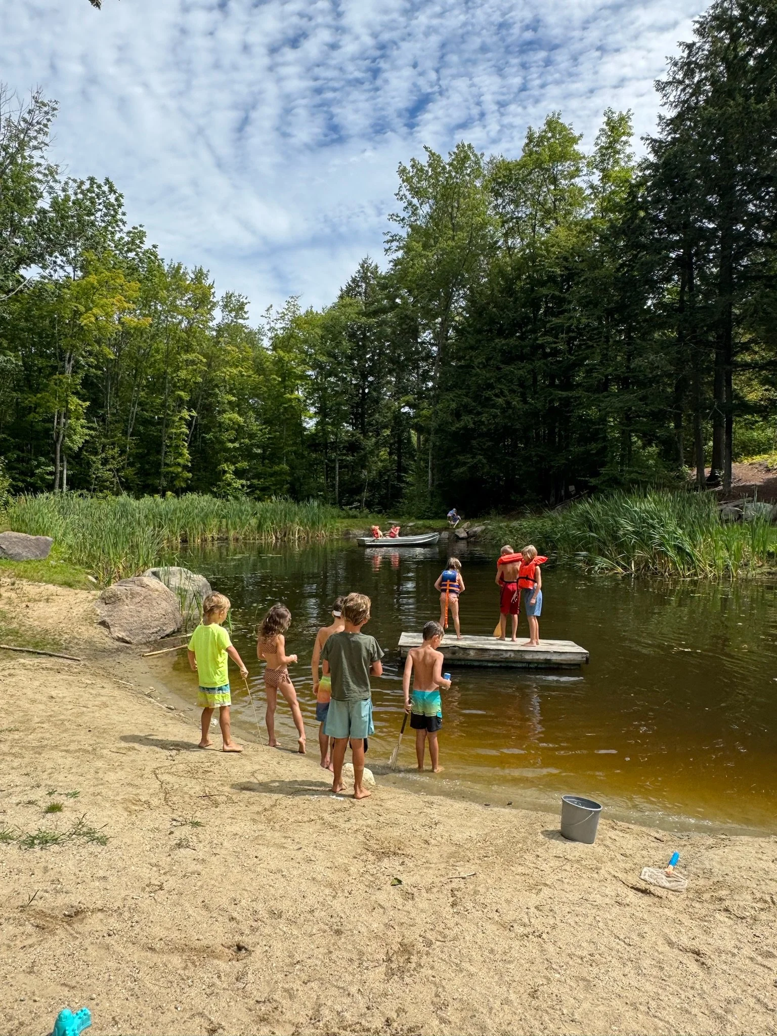 Children playing at a beach by a lake with trees and a cloudy sky in the background.