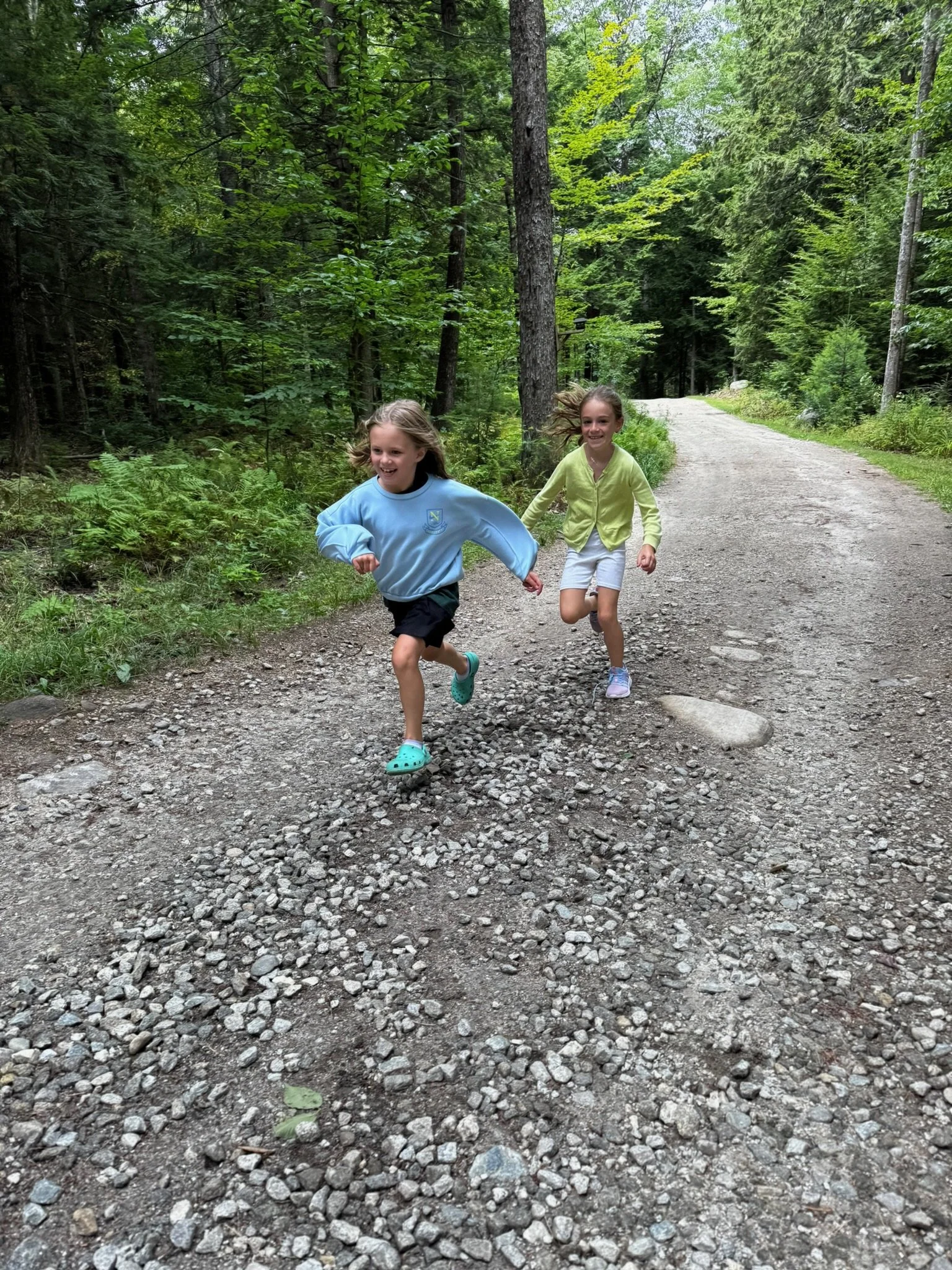 Two young girls running joyfully on a gravel trail through a green forest.
