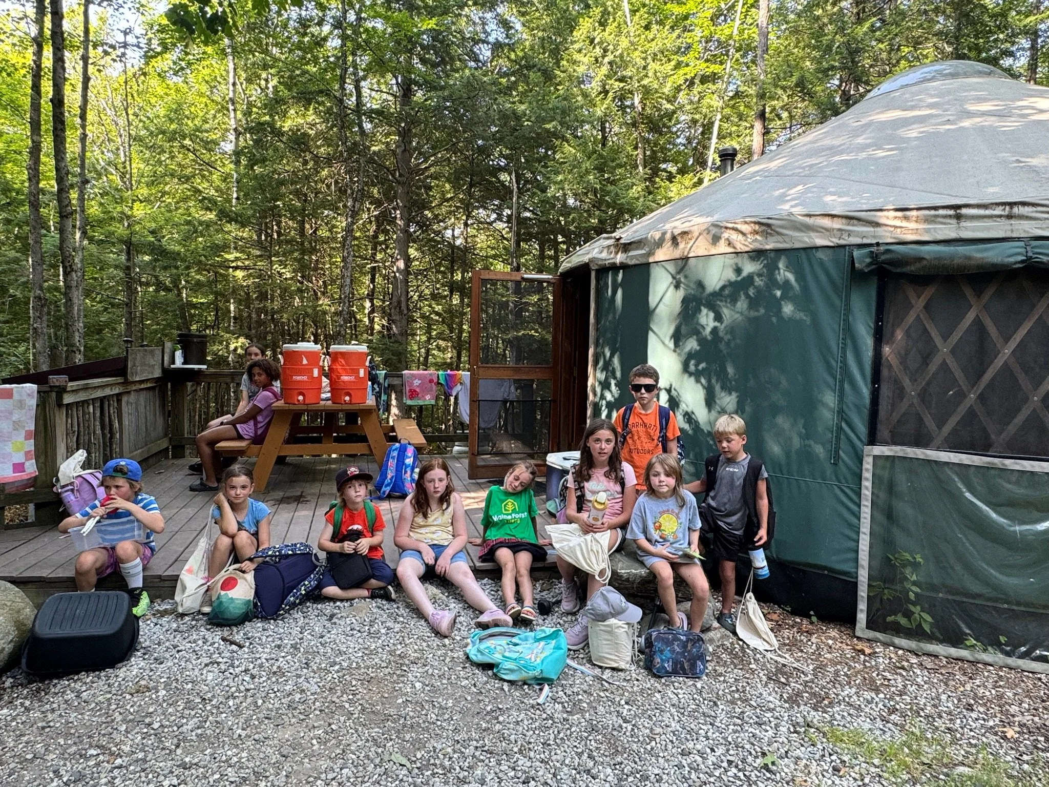 Group of children sitting and standing in front of a large green camping tent in a wooded area, with backpacks and supplies on the ground.