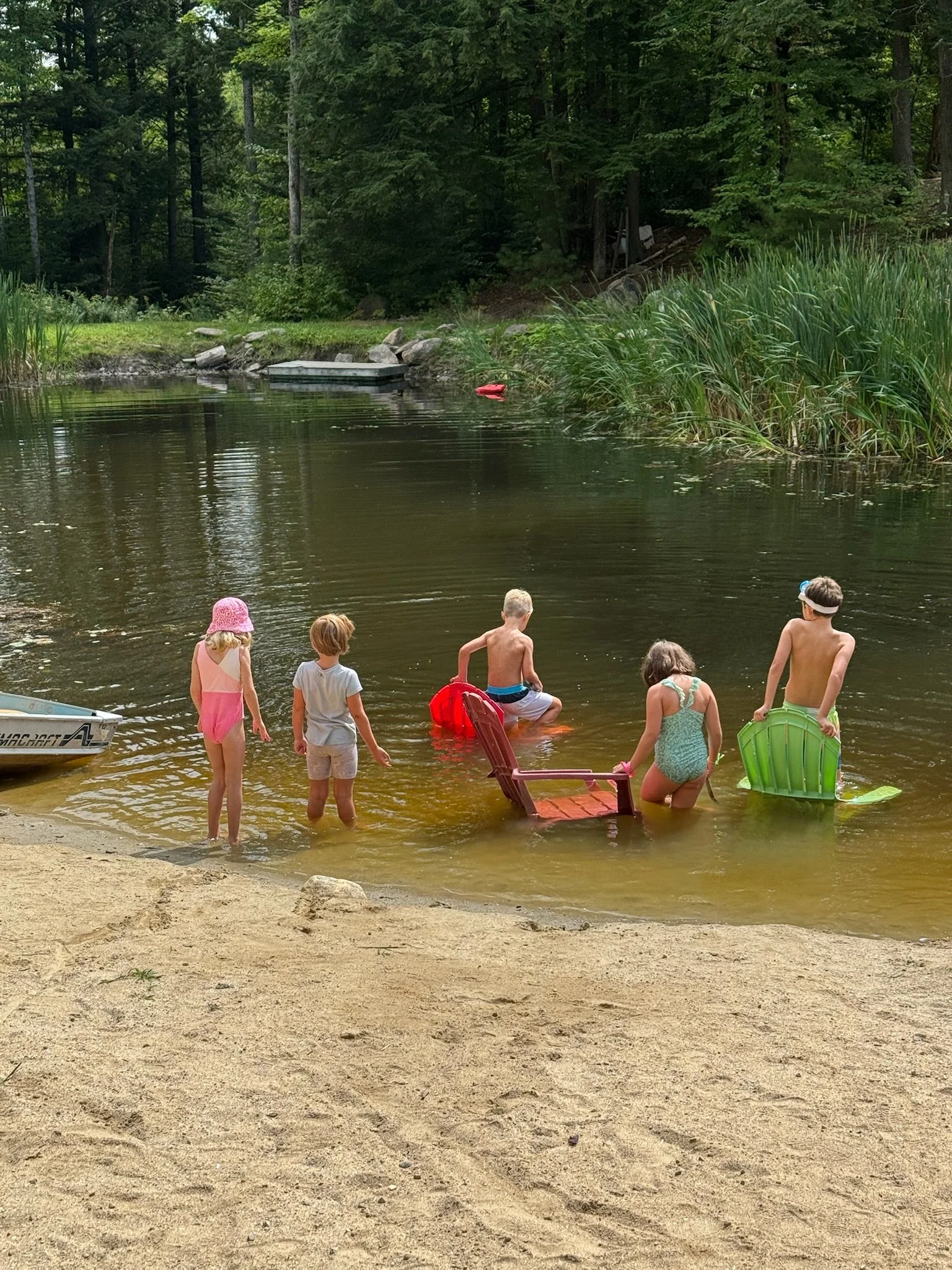 Group of six children playing and wading in a lake at a sandy shoreline surrounded by trees.
