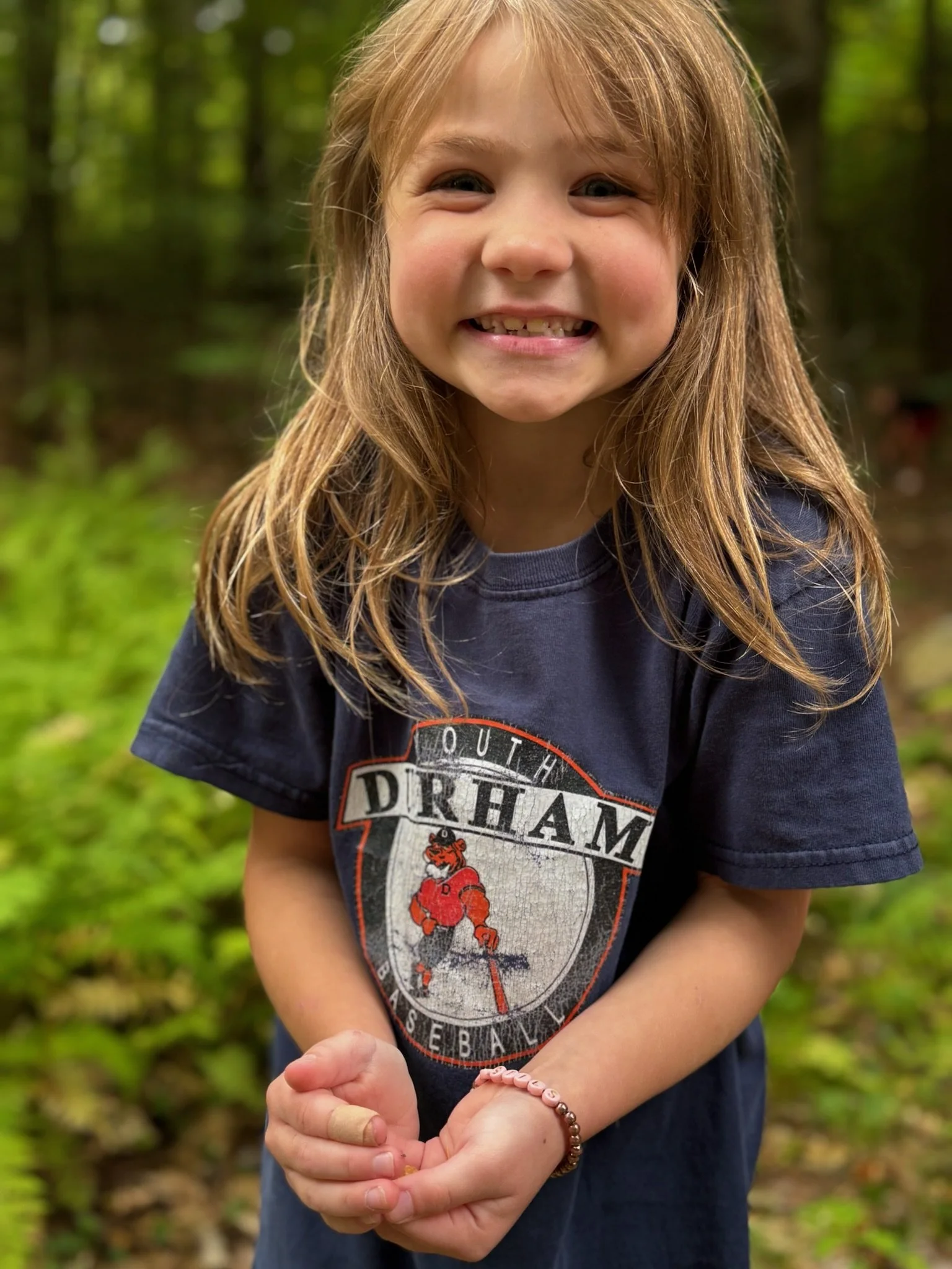 A young girl with long, light brown hair smiling outdoors in a forest, wearing a navy T-shirt with a Durham basketball logo, and clasping her hands.