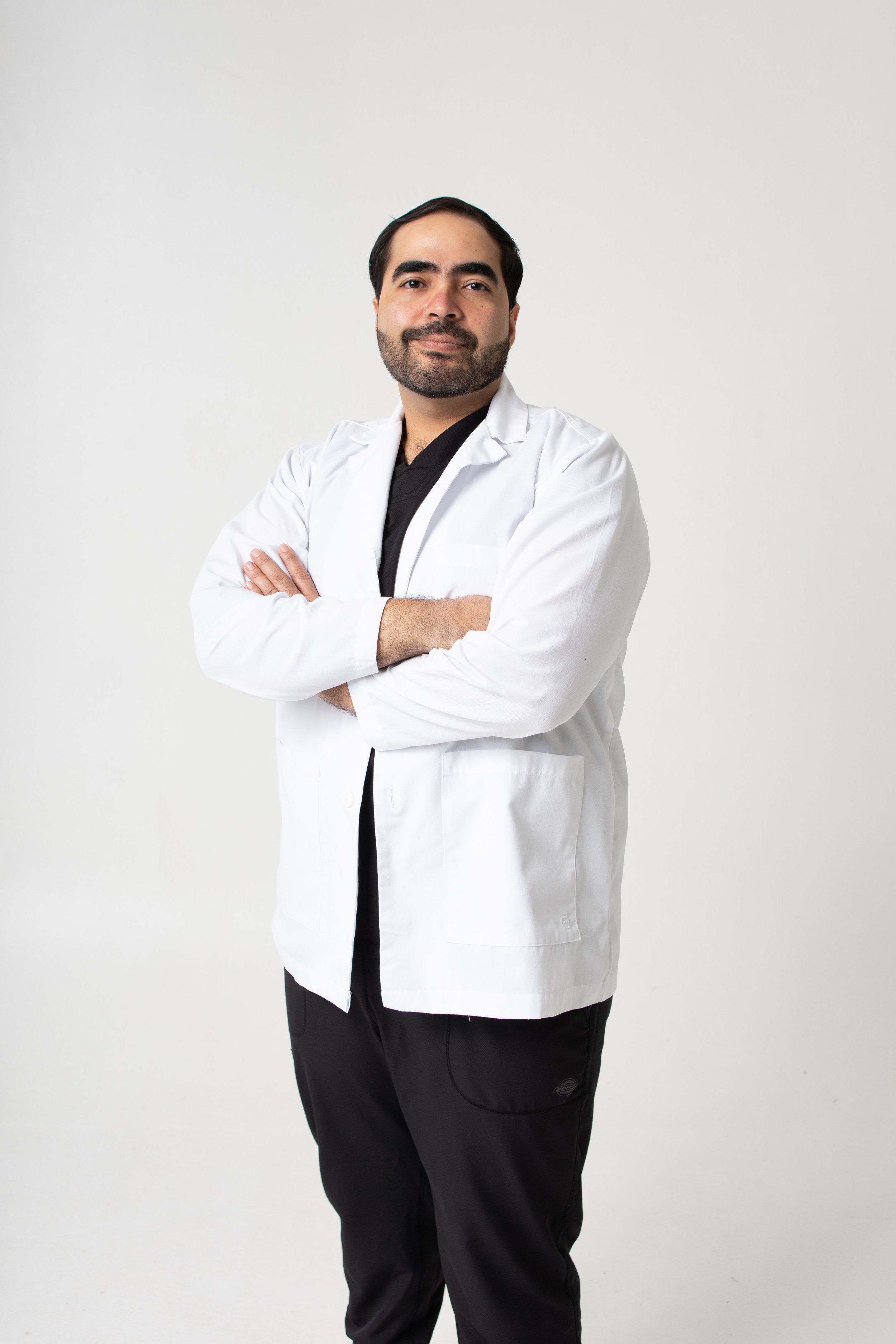 A confident male doctor with dark hair, a beard, and light skin, standing with arms crossed, wearing a white lab coat over black scrubs against a plain white background.