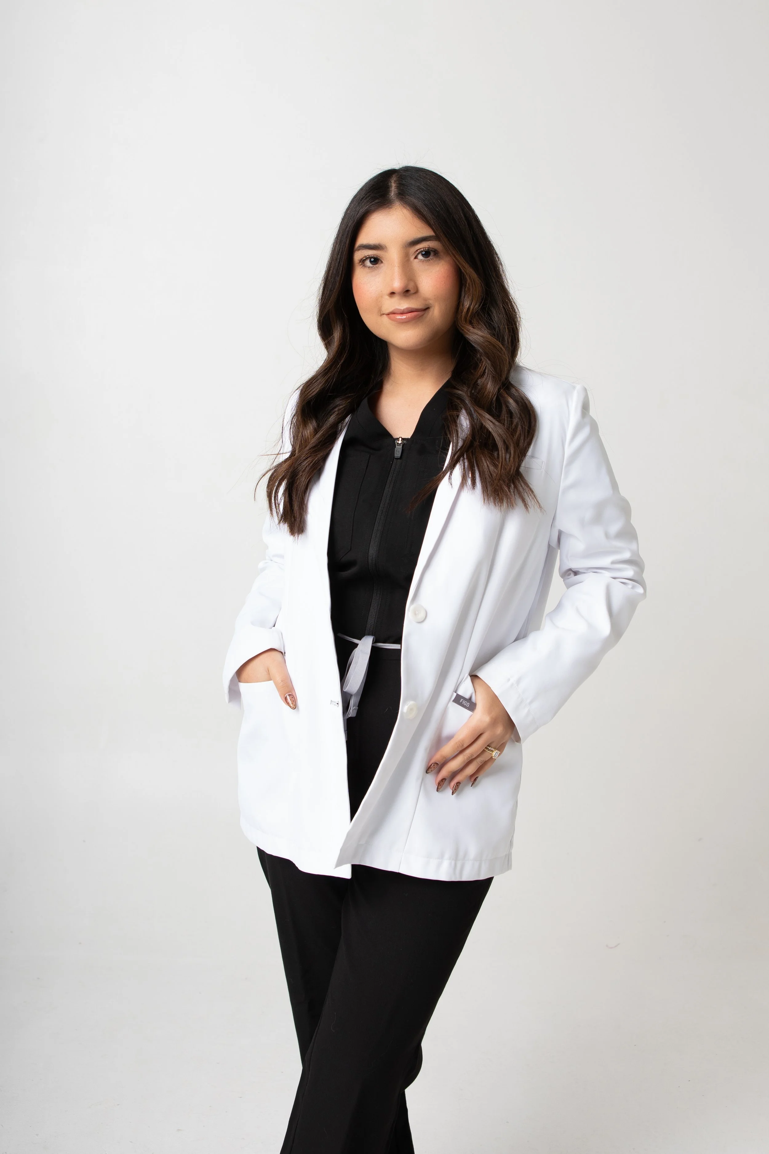 A young woman with long dark hair, wearing a white coat over black clothing, standing against a plain white background.