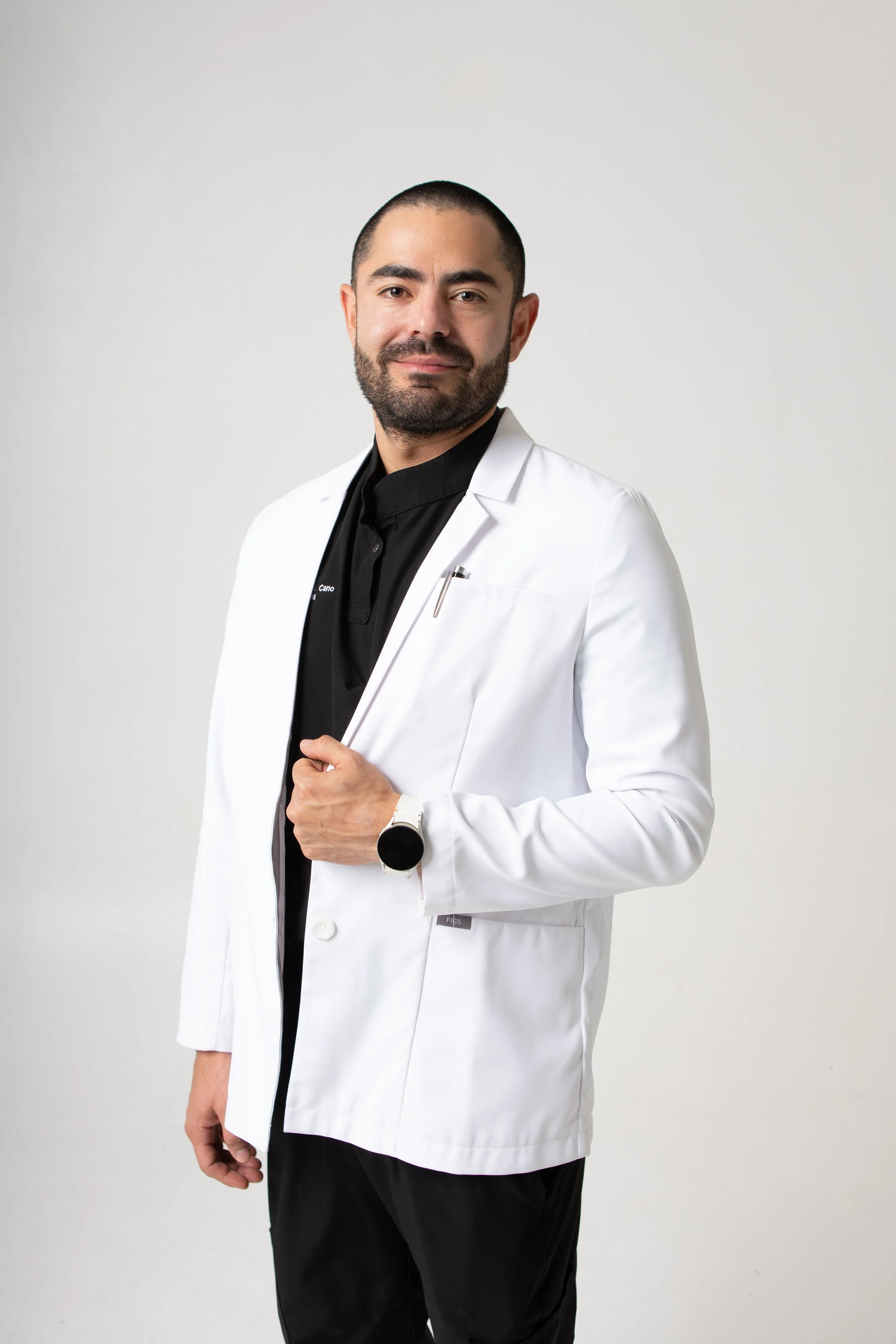 A male doctor with a beard wearing a white lab coat and a black shirt, standing against a plain white background.
