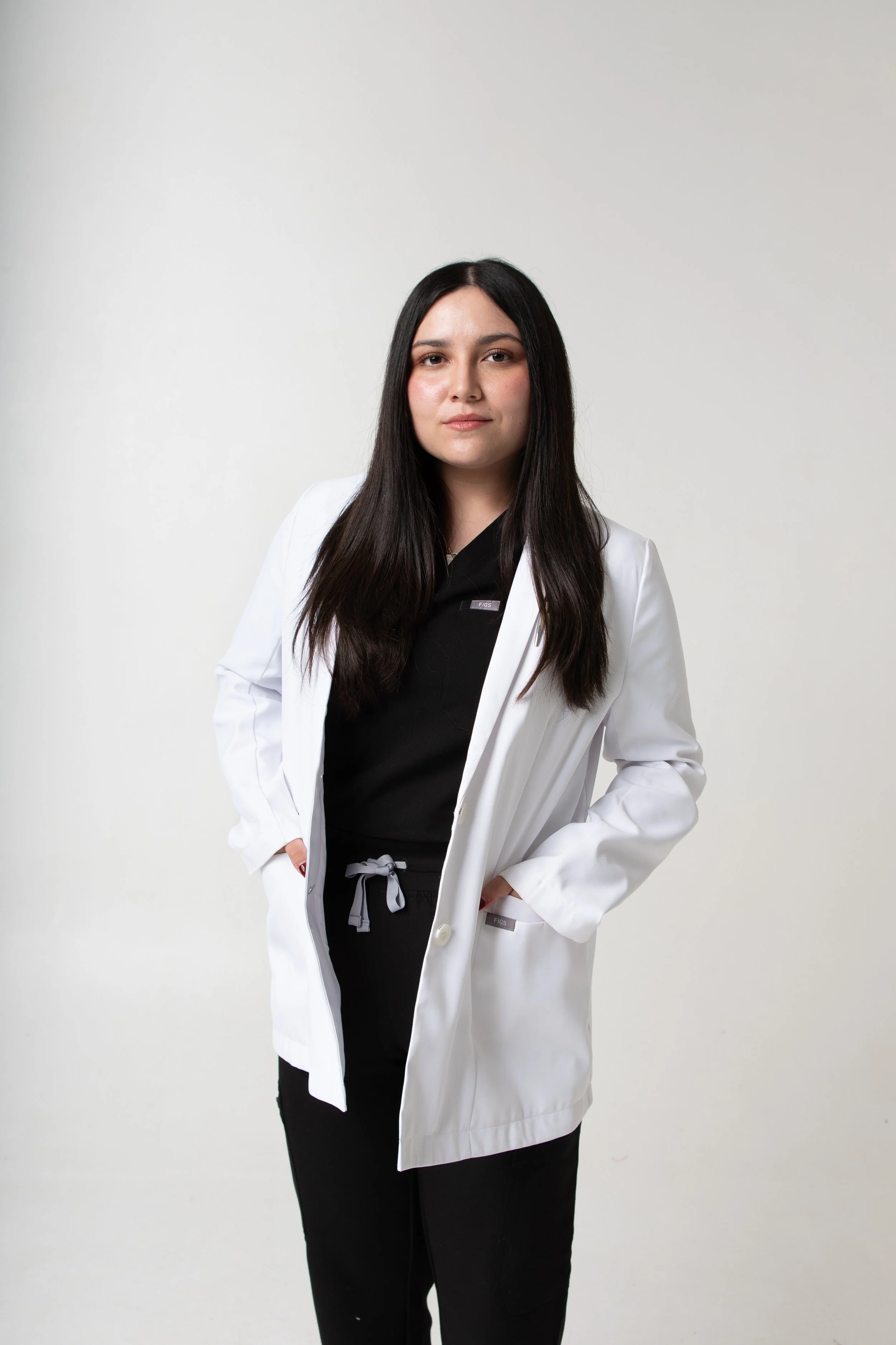 Young woman with long dark hair wearing a white lab coat over black clothing, standing with hands in pockets on a plain light background.