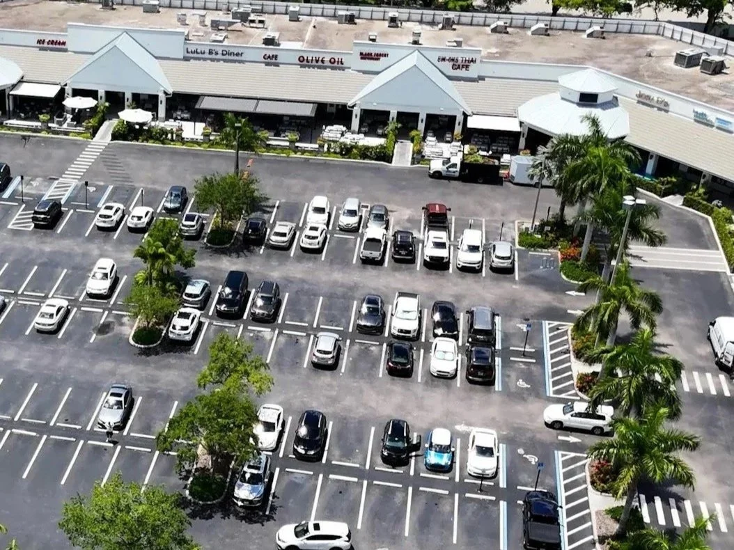 Aerial view of a shopping center parking lot with multiple parked cars and surrounding palm trees. The shopping center has several storefronts, including Lulu B's Diner and Olive Oil, with a few trucks parked in front.