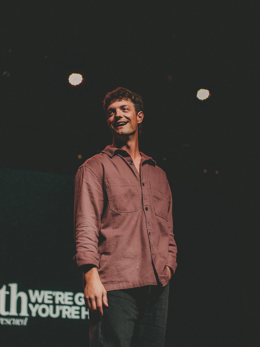A young man in a brown shirt smiling and standing on a stage with lights above and a black background.
