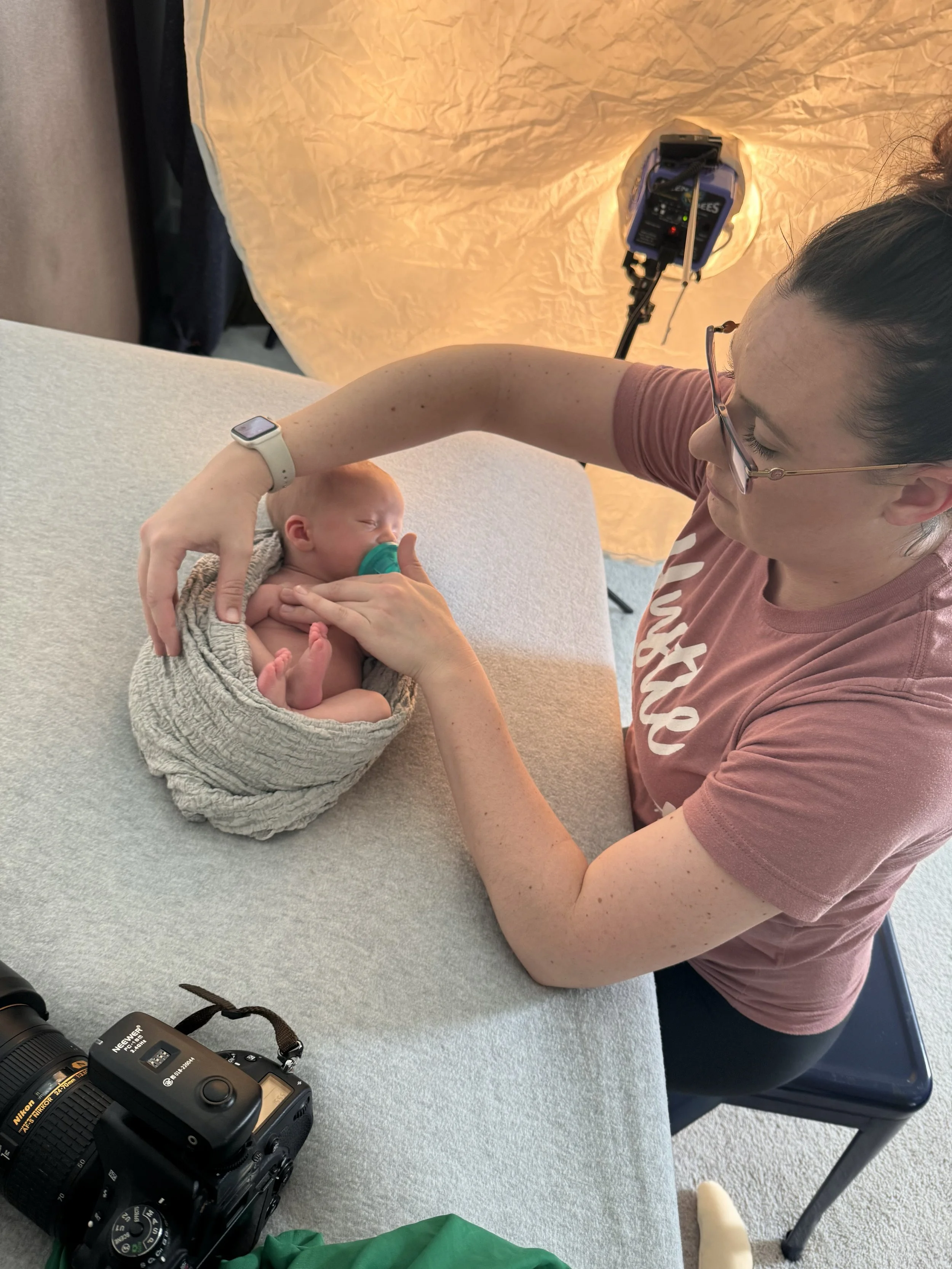 A woman is gently holding a newborn baby on a table during a photo shoot. The baby is wrapped in a cozy blanket and has a pacifier. The woman is adjusting the baby's position, with a camera and studio lighting setup visible in the background.