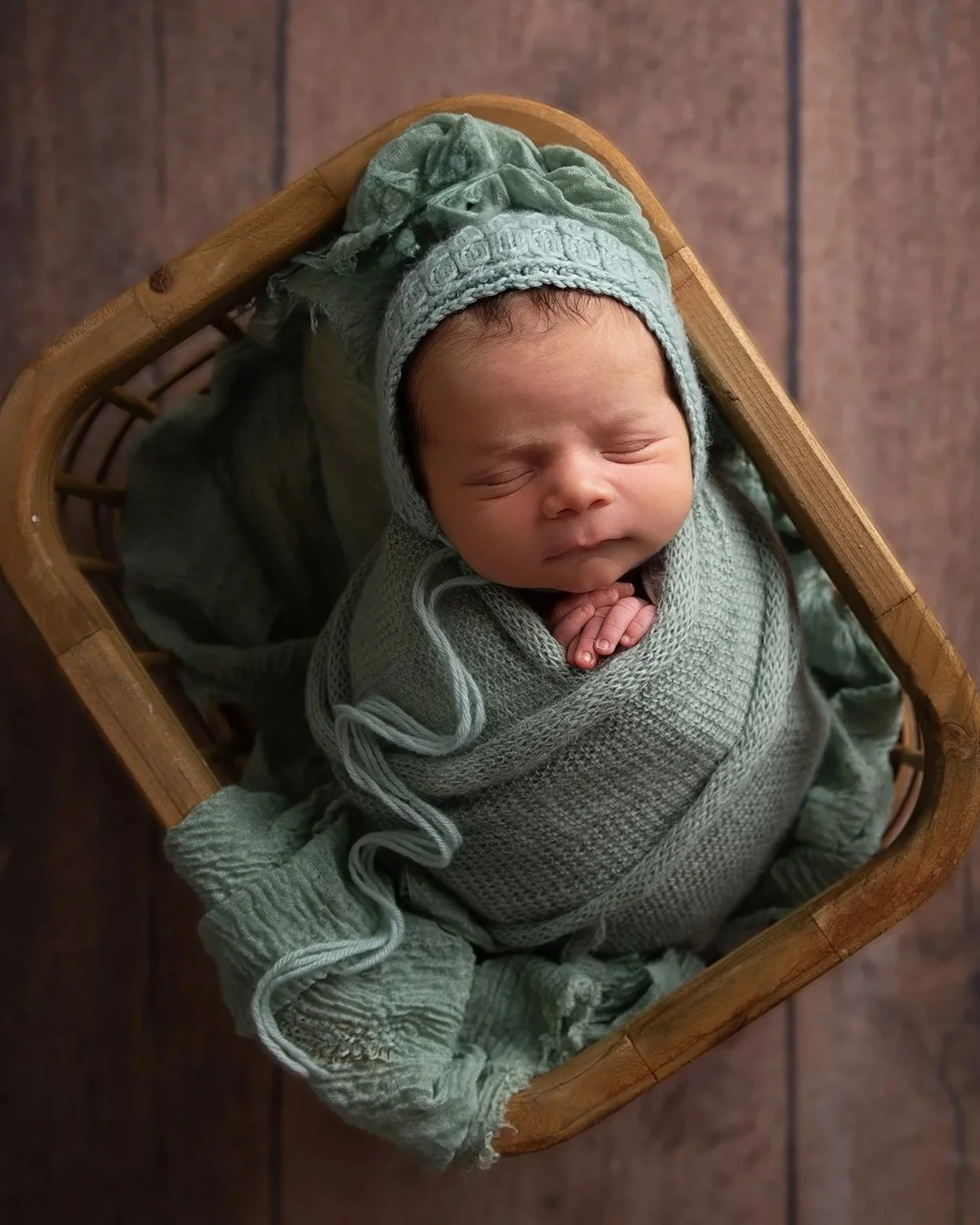 A newborn baby peacefully sleeping, swaddled in a green knitted blanket, wearing a matching bonnet, inside a wooden basket.