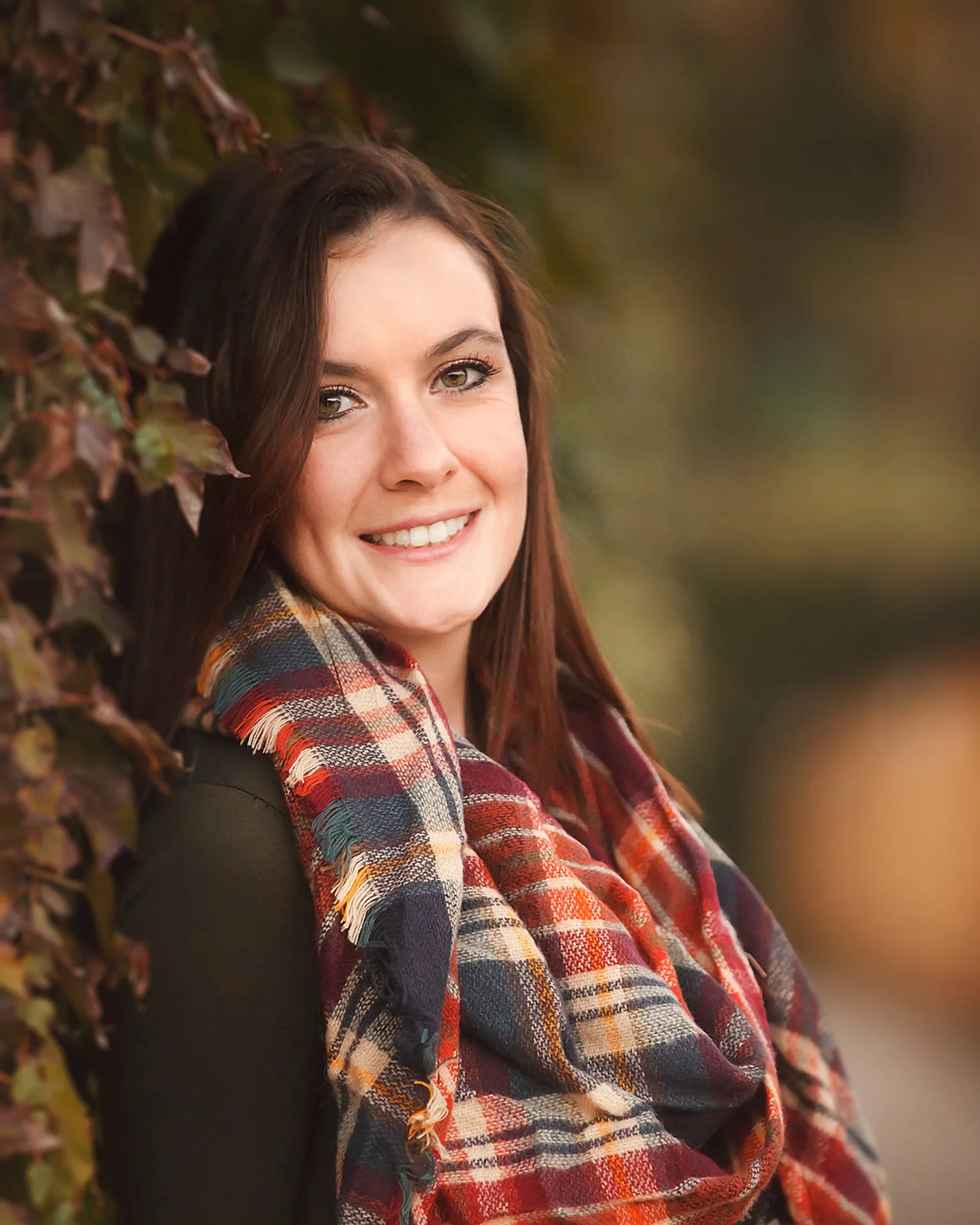 A young woman with brown hair smiling outdoors, wearing a black top and a colorful plaid scarf.