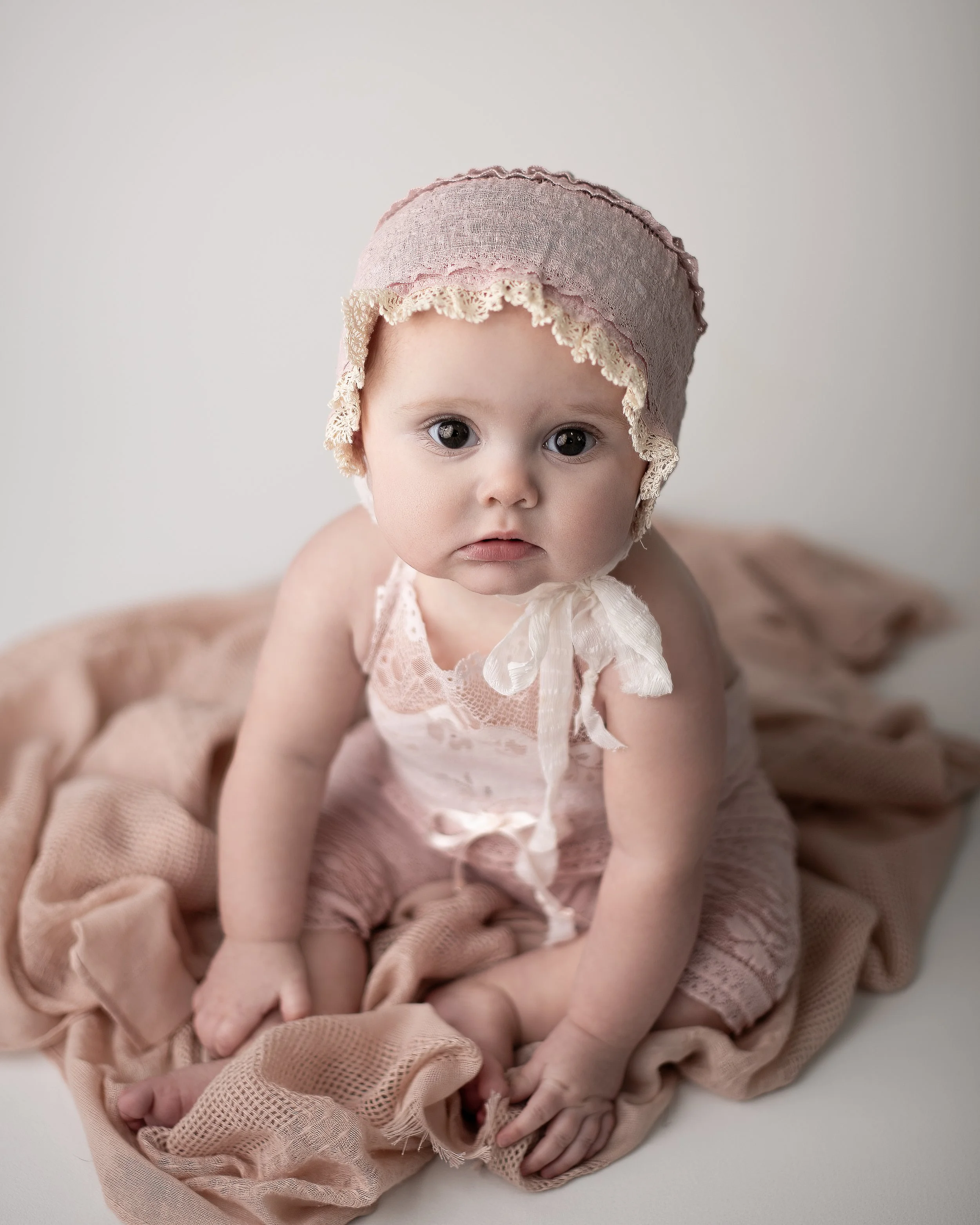 A baby girl with big eyes wearing a pink bonnet with lace, sitting on a soft pink blanket, looking at the camera.