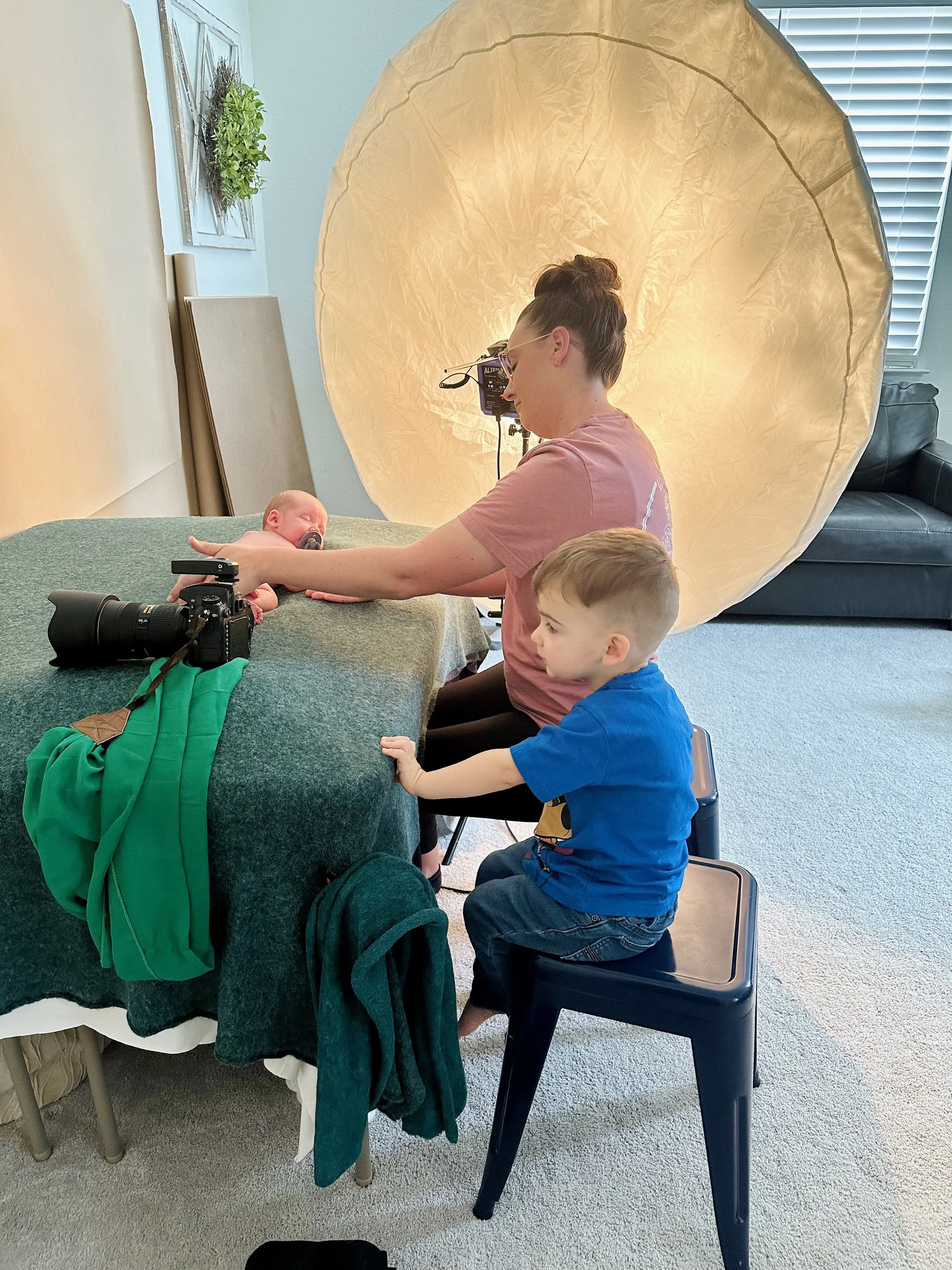 A woman and a young boy are taking a photograph of a newborn baby with a digital camera in a cozy room. The woman is sitting at a table with a large, circular light diffuser behind her, and the boy is sitting on a small black stool, holding onto the woman's arm as she adjusts her position to photograph the baby.